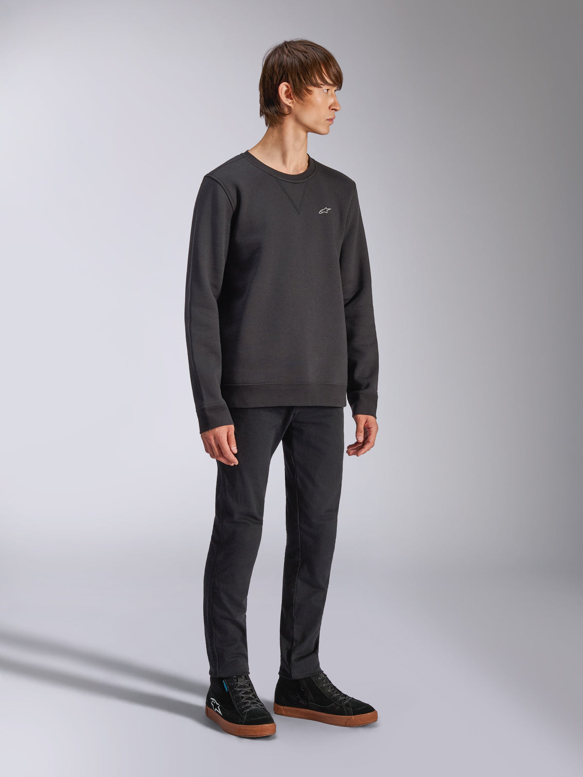 Alpinestars Positive Crew, Pullover Sweatshirt, Black, small white Alpinestars logo on the chest, ribbed collar with V-stitch detail, ribbed cuffs and hem, worn by a model with black trousers and sneakers, casual motorsport-inspired streetwear