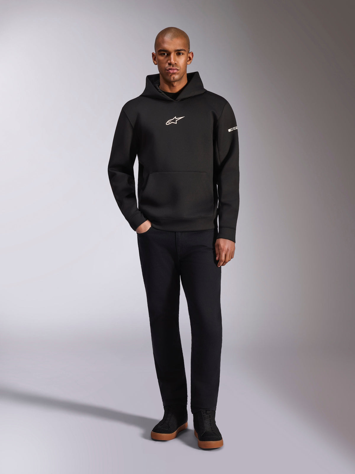 Alpinestars Insight Hoodie, black pullover hoodie with white Astars logo on the chest and sleeve, worn by a model with black pants and black sneakers, casual motorsport-inspired streetwear