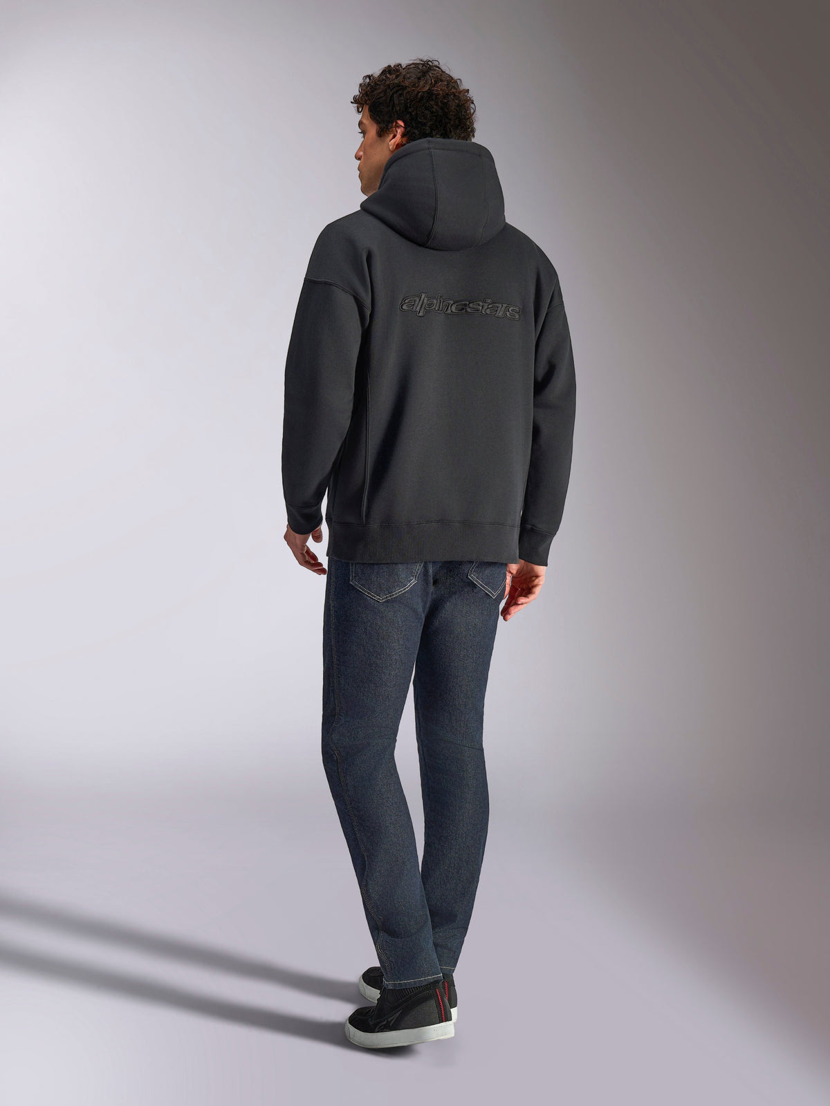 Alpinestars Emergent Oversized Hoodie, Black, back view showing 3D embroidered Alpinestars word logo across the shoulders, oversized fit, styled with dark blue denim jeans and black sneakers, casual motorsport-inspired streetwear.