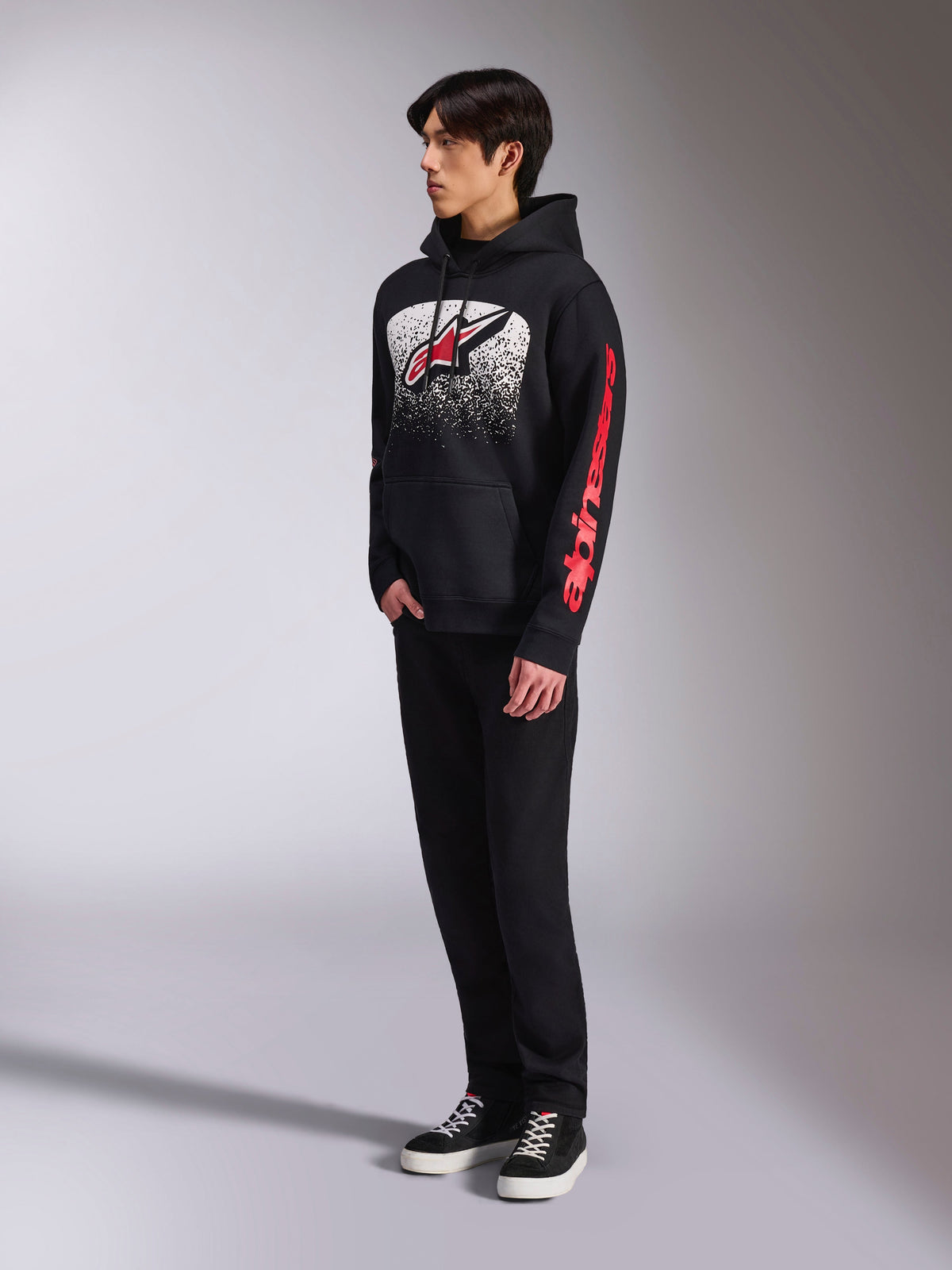 Alpinestars Obsessed Hoodie, black pullover fleece, featuring a large white speckled chest graphic with a red Astars logo and red sleeve branding, worn by a model with black pants and sneakers, casual motorsport streetwear