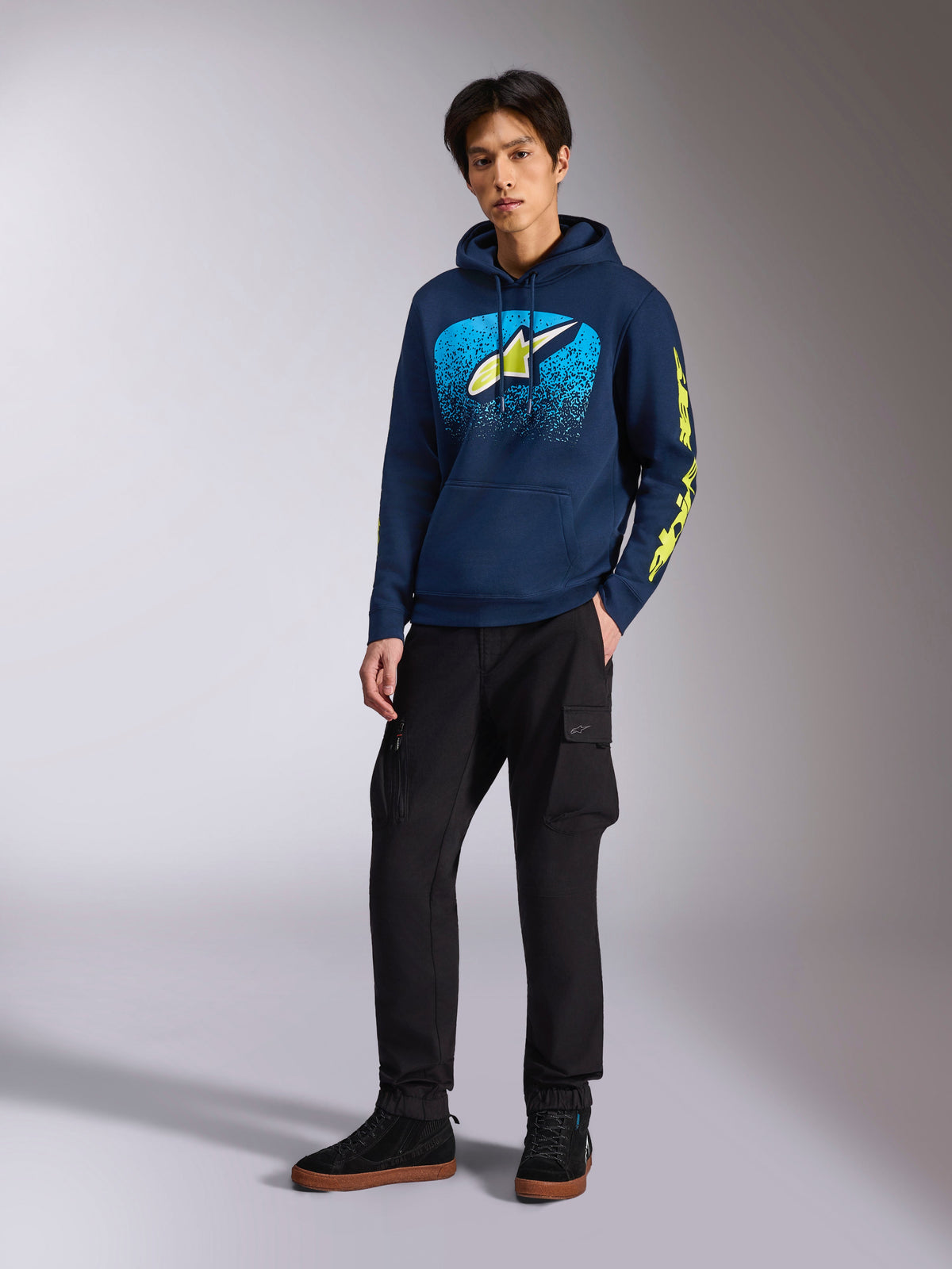 Alpinestars Obsessed Hoodie in Navy, casual pullover fleece with light blue and lime yellow front graphic, lime yellow Alpinestars sleeve branding, worn by a model with black cargo pants and black sneakers, motorsport-inspired streetwear