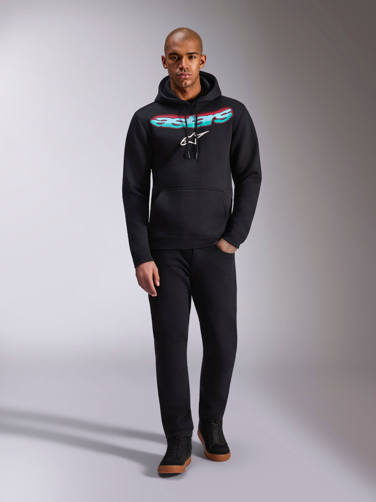 Alpinestars Ellipsoid Hoodie, Black, Pullover fleece featuring a curved Astars wordmark with a red and teal color gradient and white star logo, worn by model with black pants and sneakers, casual motorsport streetwear