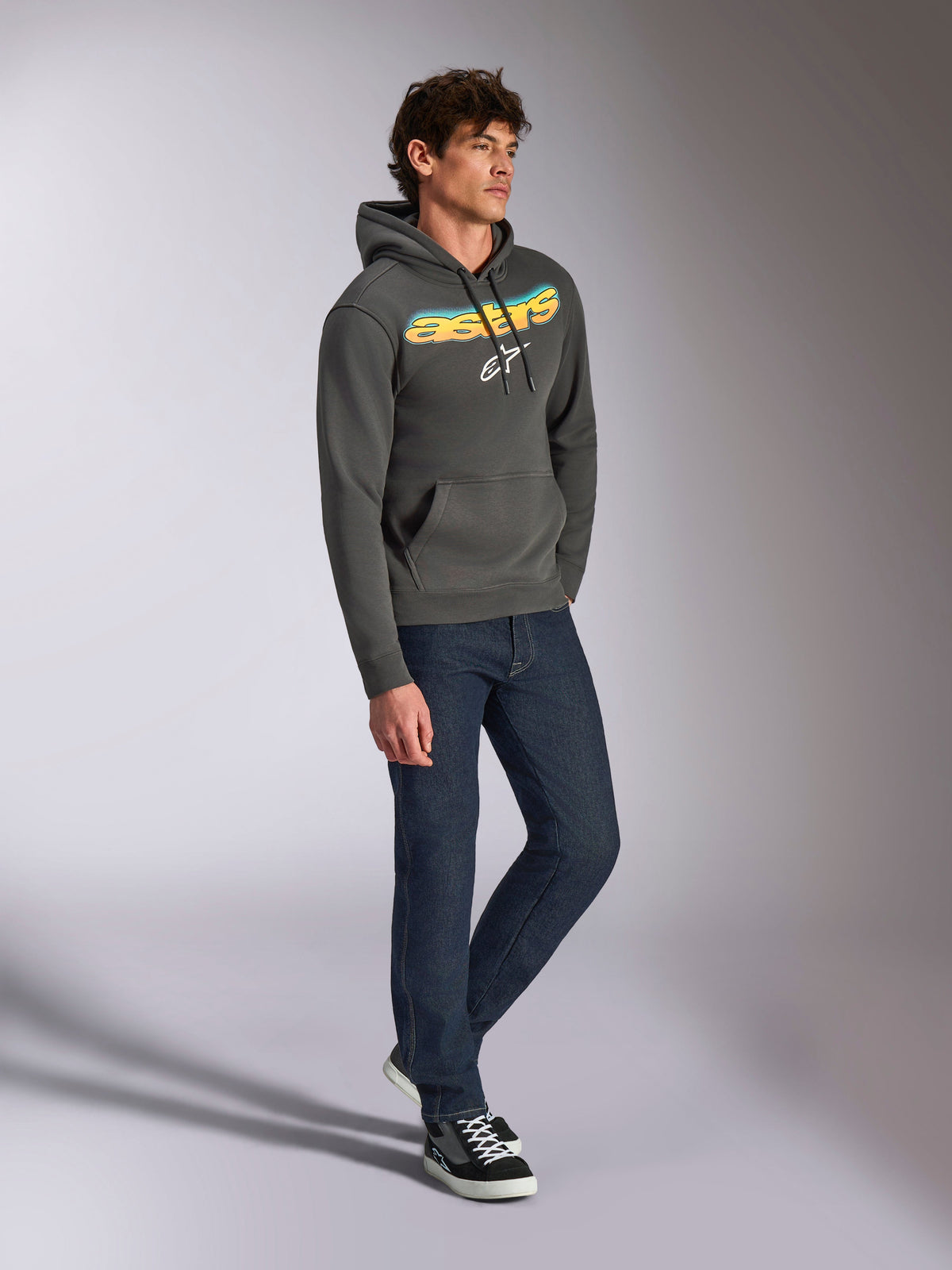Alpinestars Ellipsoid Hoodie, Gunmetal gray pullover fleece, front curved Astars wordmark with blue and yellow color gradient and white logo, styled with dark jeans and black sneakers, casual streetwear.