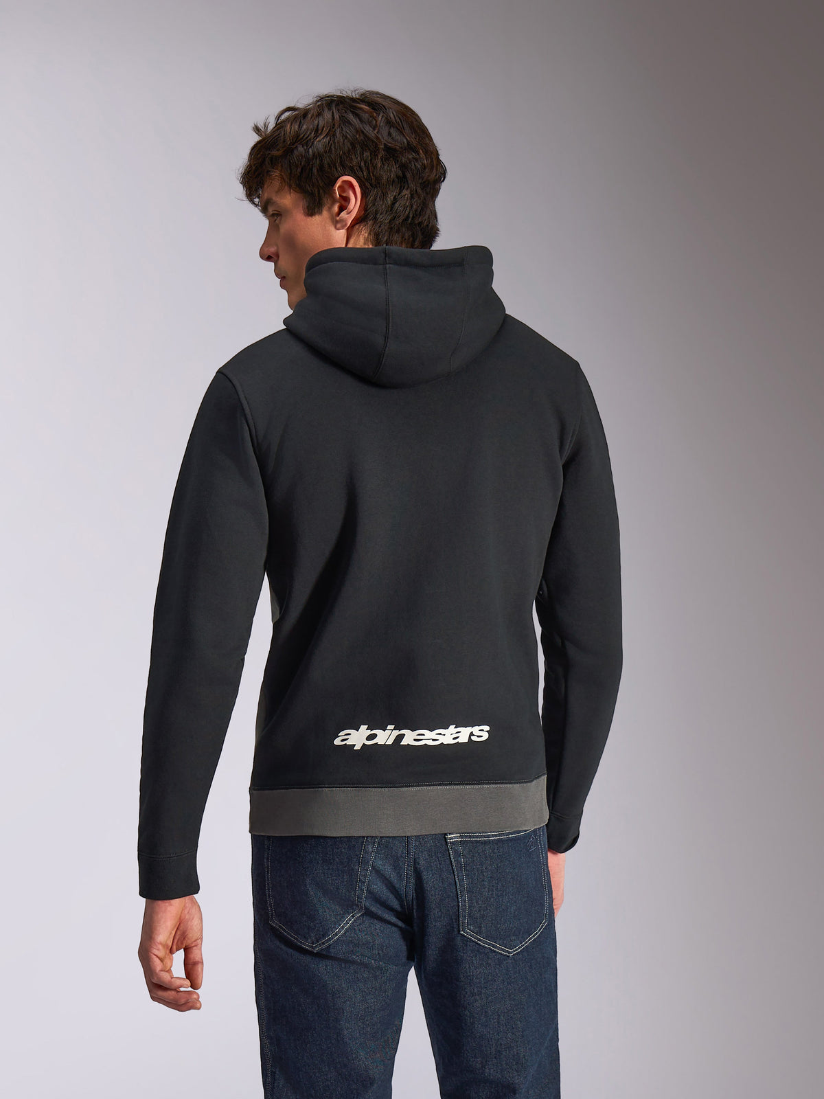Alpinestars Parallel Hoodie, casual sportswear hoodie, Black and Gunmetal, back view featuring a white Alpinestars logo on the lower back, color-blocked design with gray hem, worn with dark denim jeans