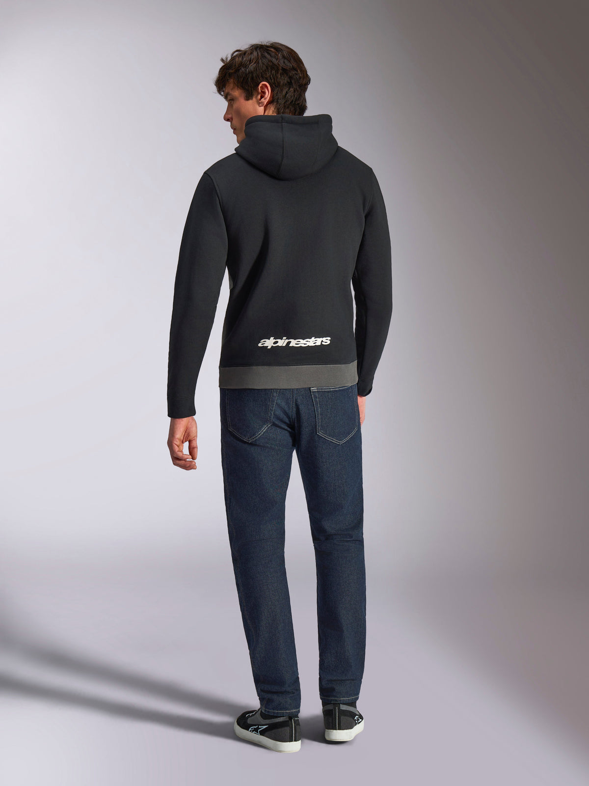 Alpinestars Parallel Hoodie, rear view of a man wearing a black and gunmetal color-blocked pullover fleece, white Alpinestars word logo on the lower back, paired with dark blue jeans, casual motorsport-inspired streetwear
