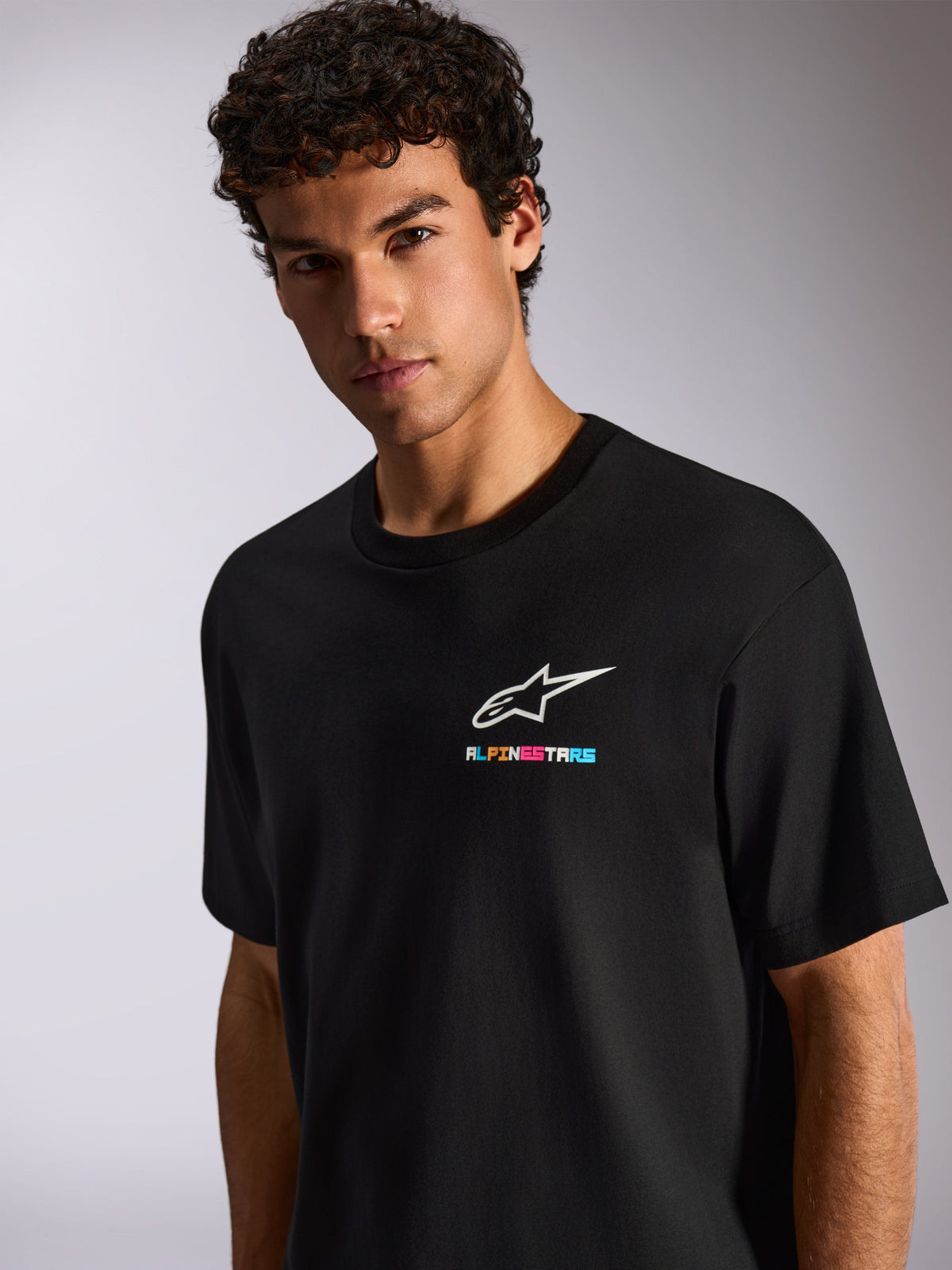 A man wearing the Alpinestars Wild Oversized CSF Tee, Black Short Sleeve T-Shirt, featuring a white Astars logo and vibrant multi-colored branding on the chest, casual motorsport streetwear