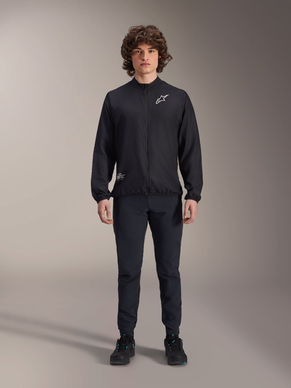 Model wearing Alpinestars A-Aria Windbreaker Jacket in Black, featuring a full-zip front, mock neck, and white Astars logo on the chest, paired with black technical pants and black shoes, performance cycling apparel for mountain biking and outdoor use.