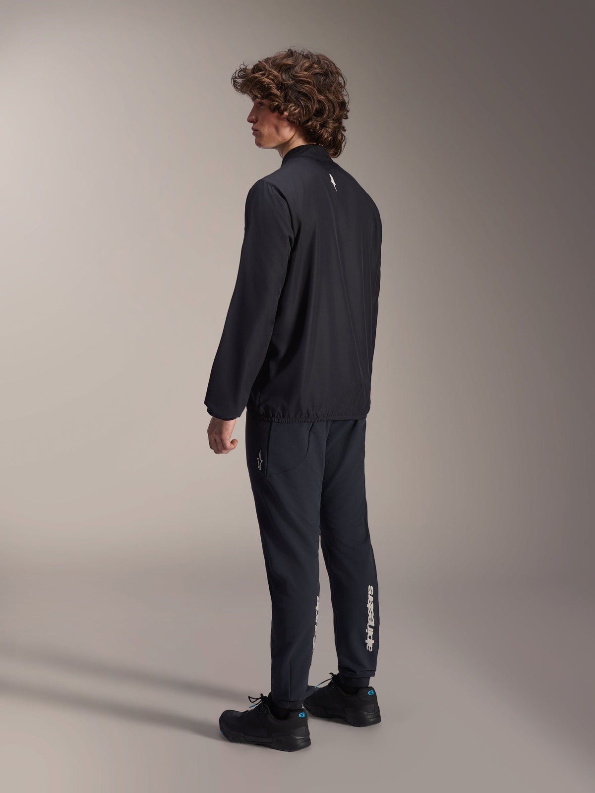 Alpinestars A-Aria Windbreaker Jacket, Black, lightweight wind-resistant cycling jacket, rear three-quarter view on model, featuring a small white logo on the back neck and elasticated hem, paired with matching black pants and shoes for a performance-driven look.