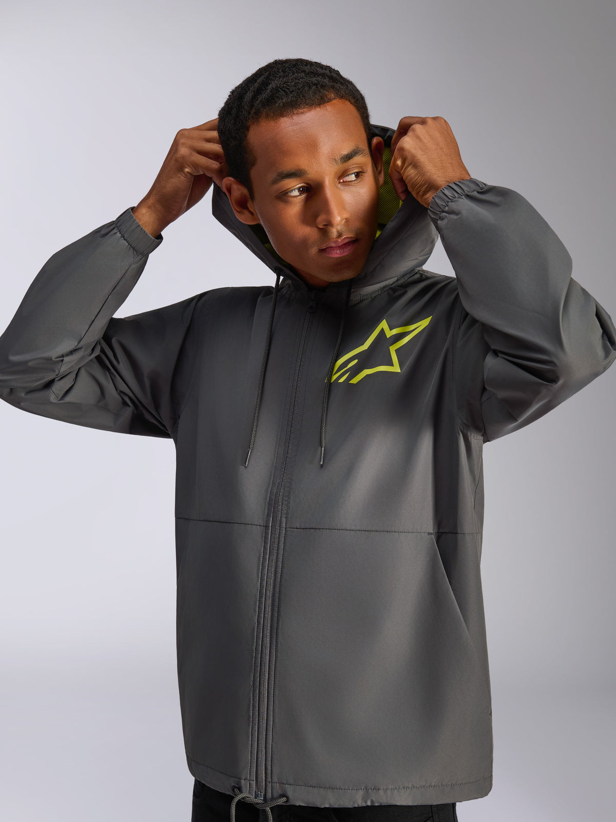 Alpinestars Speeder Chromium Windbreak Jacket, Charcoal Grey and Hi-Vis Yellow, male model wearing hooded windbreaker with drawstring, hi-vis yellow Alpinestars logo on chest and yellow hood lining, casual motorsport-inspired streetwear