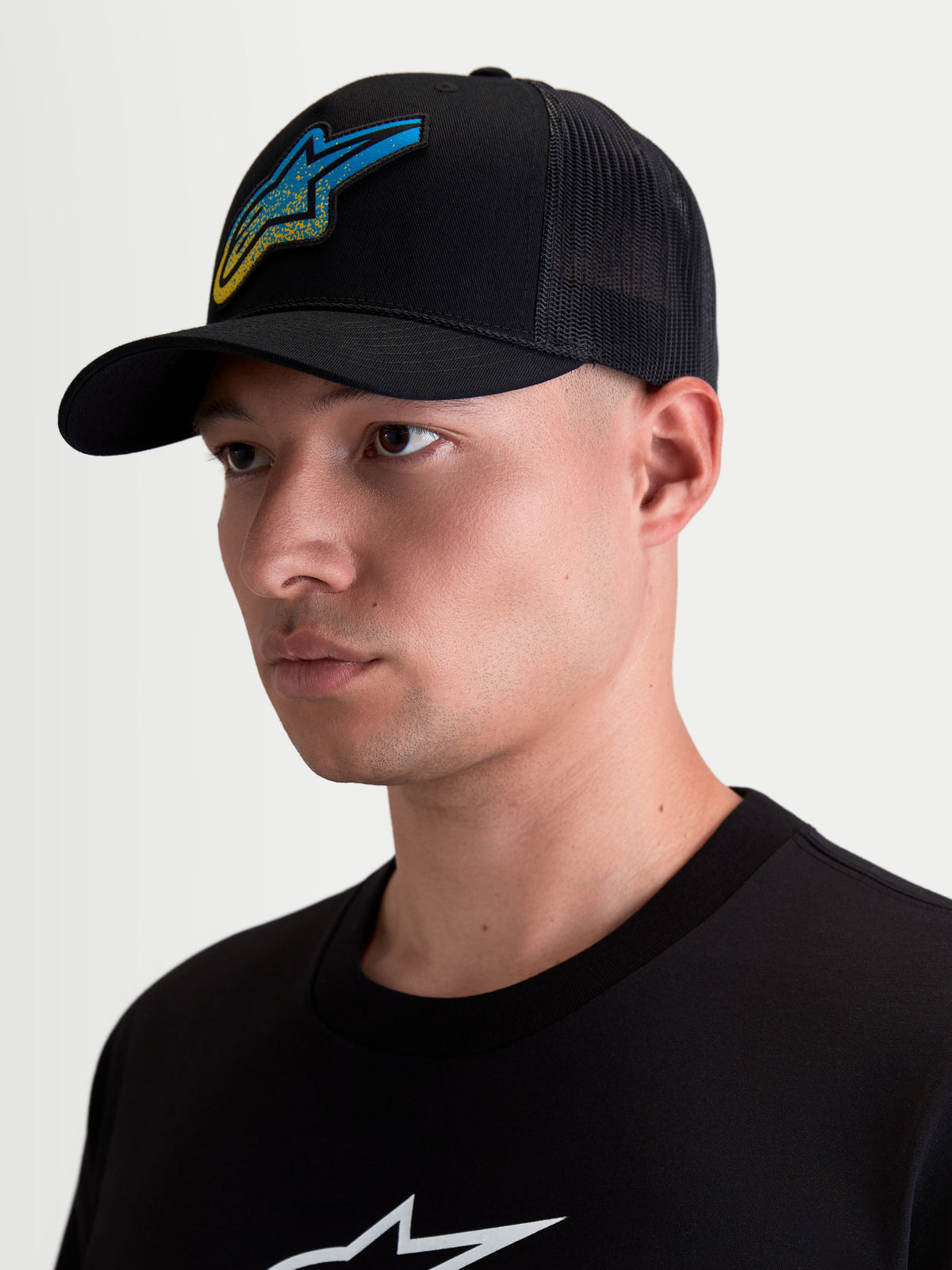 Alpinestars Meteorite Trucker Hat, Sportswear Casual Lifestyle Mesh Back Cap, Men's, Black, featuring a blue and yellow Astars gravel texture woven logo patch, curved bill, and structured crown, shown on model.