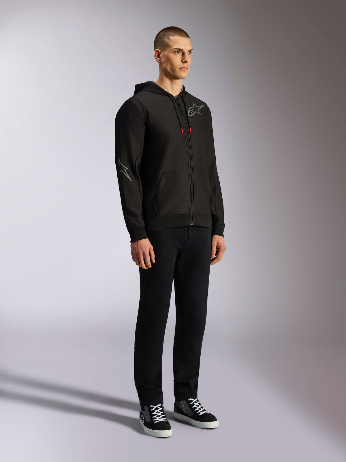 Alpinestars Sessions V3 Hoodie, casual motorsport streetwear, black zip-up hoodie with gray star logo on chest and sleeve, red-tipped drawcords, rib cuffs and hem, worn by model with black pants and sneakers.