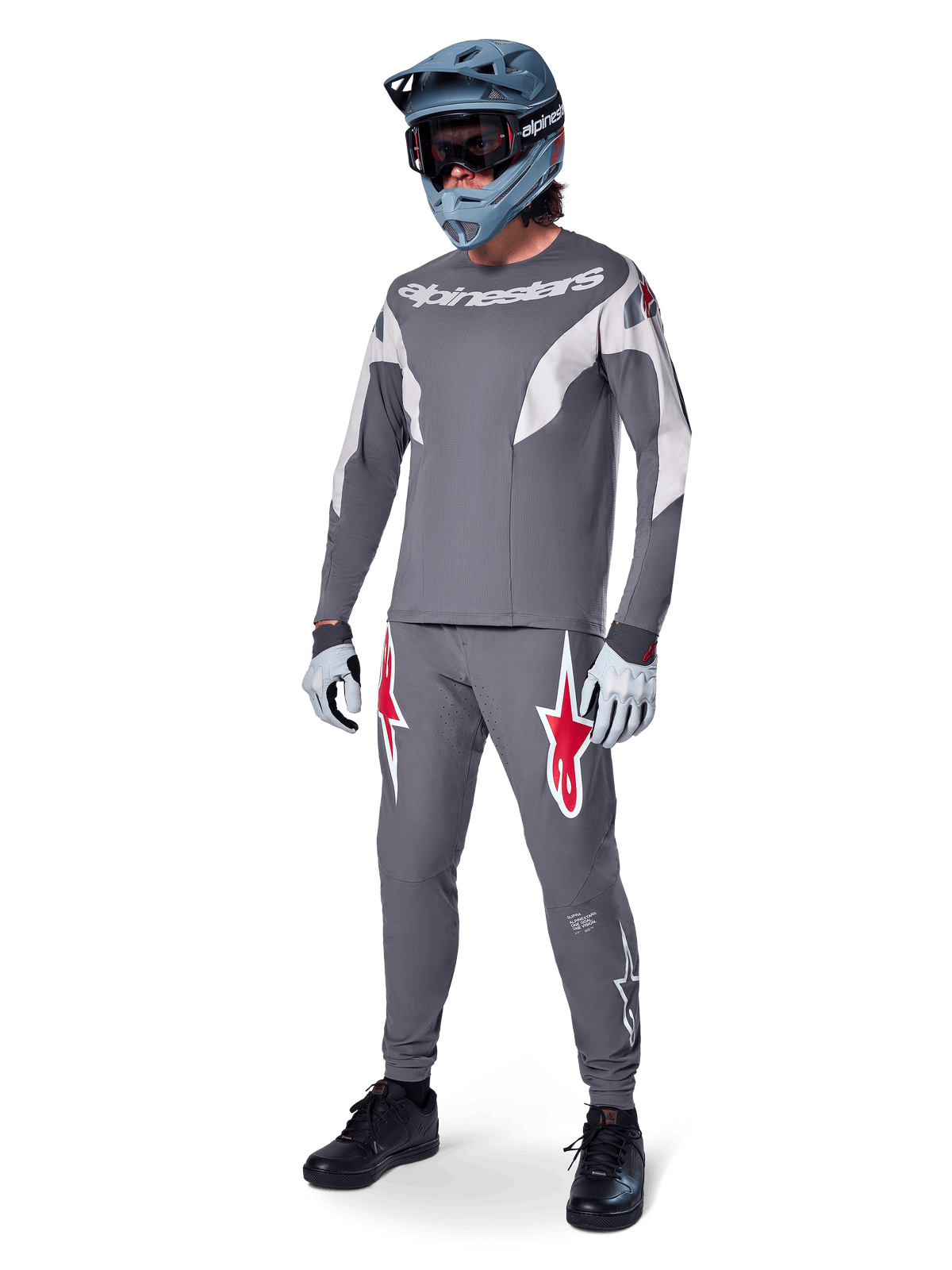 Alpinestars A-Supra Race Pants in Dark Gray, featuring bold red and white logos on the thighs, worn by a rider as part of a complete mountain bike racing kit with a matching gray jersey, blue-gray full-face helmet, and light gray gloves, high-performance gear for MTB racing