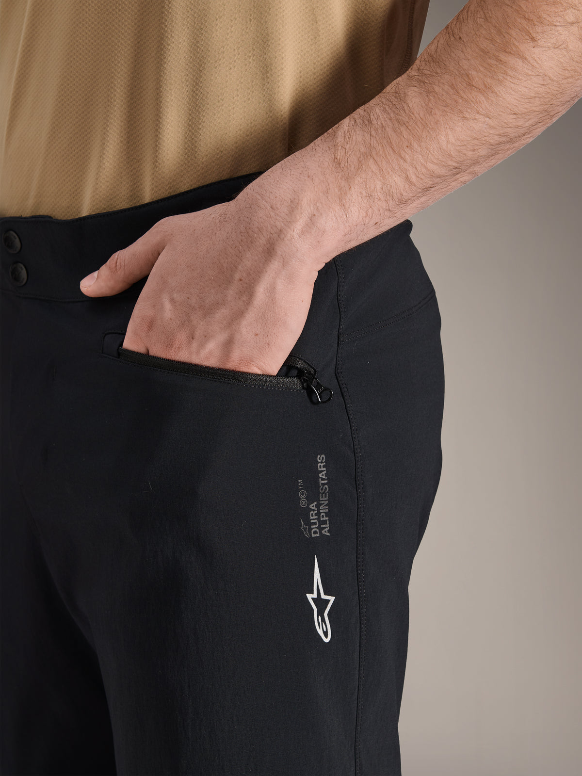 Alpinestars A-Dura Shorts, Black Mountain Bike Shorts, close-up shot of zippered side pocket with hand inside, featuring white Alpinestars logo and A-Dura branding, durable textile for off-road cycling