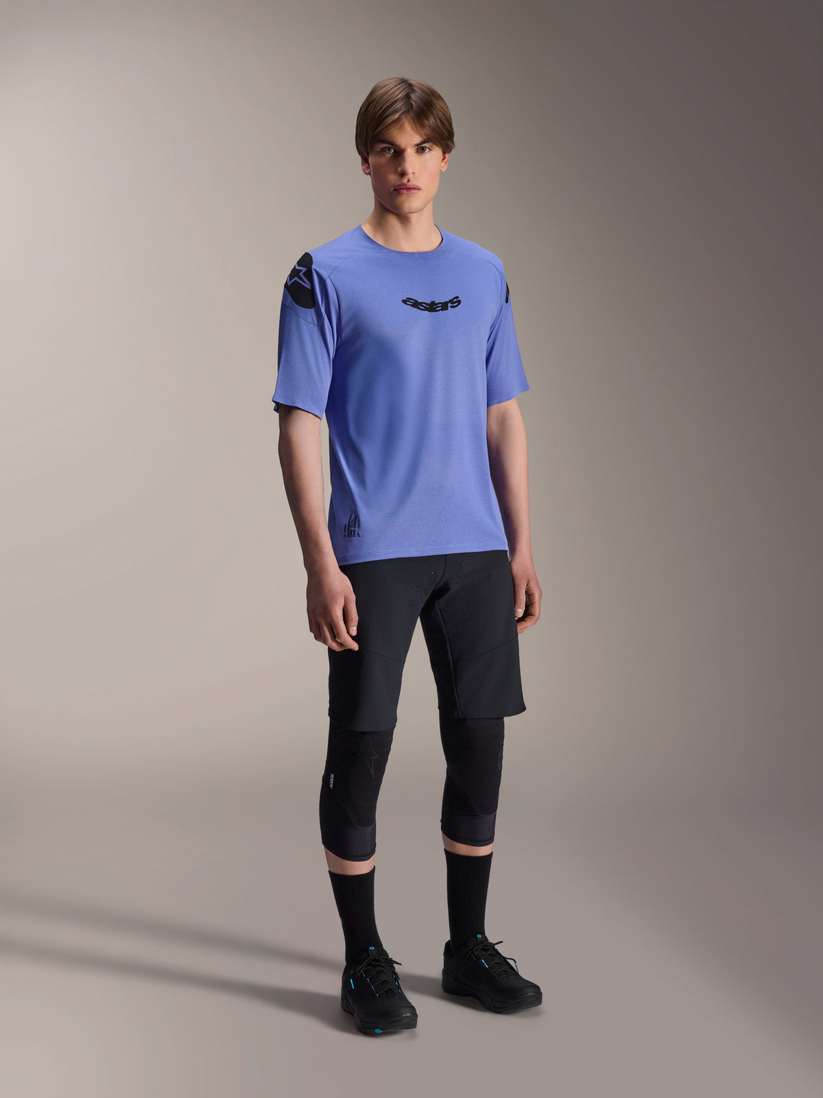 Male model wearing Alpinestars A-Aria Elite Shorts in Black Titanium and A-Aria Polartec Elite SS Jersey in light purple, paired with black mountain bike knee pads and shoes, professional cycling gear for off-road performance.