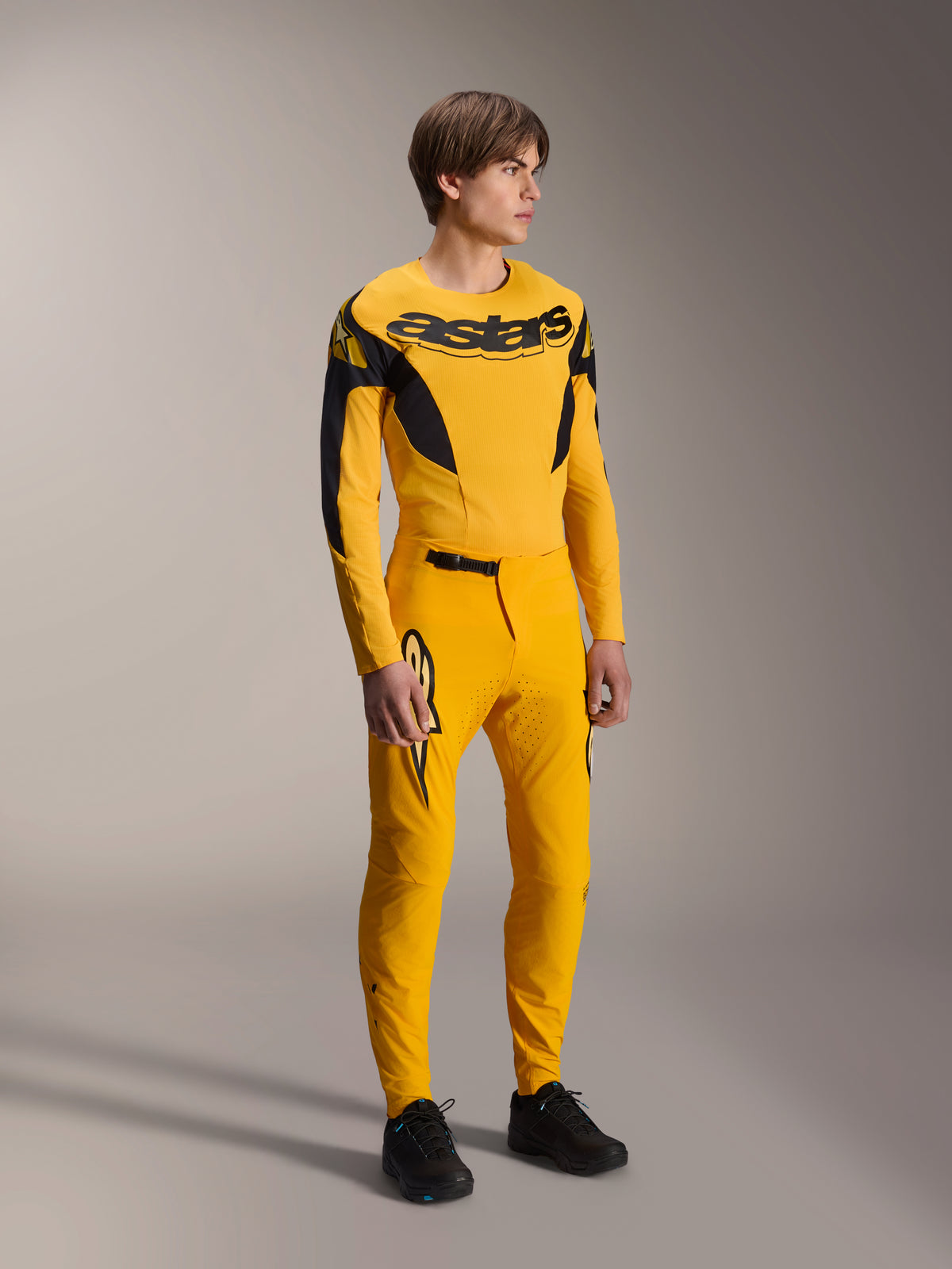 Male model wearing Alpinestars Limited Edition A-Supra Melt Gold Gear-Set, yellow long-sleeve MTB jersey with bold black branding on the chest, matching yellow pants with laser-cut ventilation and black accents, complete mountain bike racing kit.