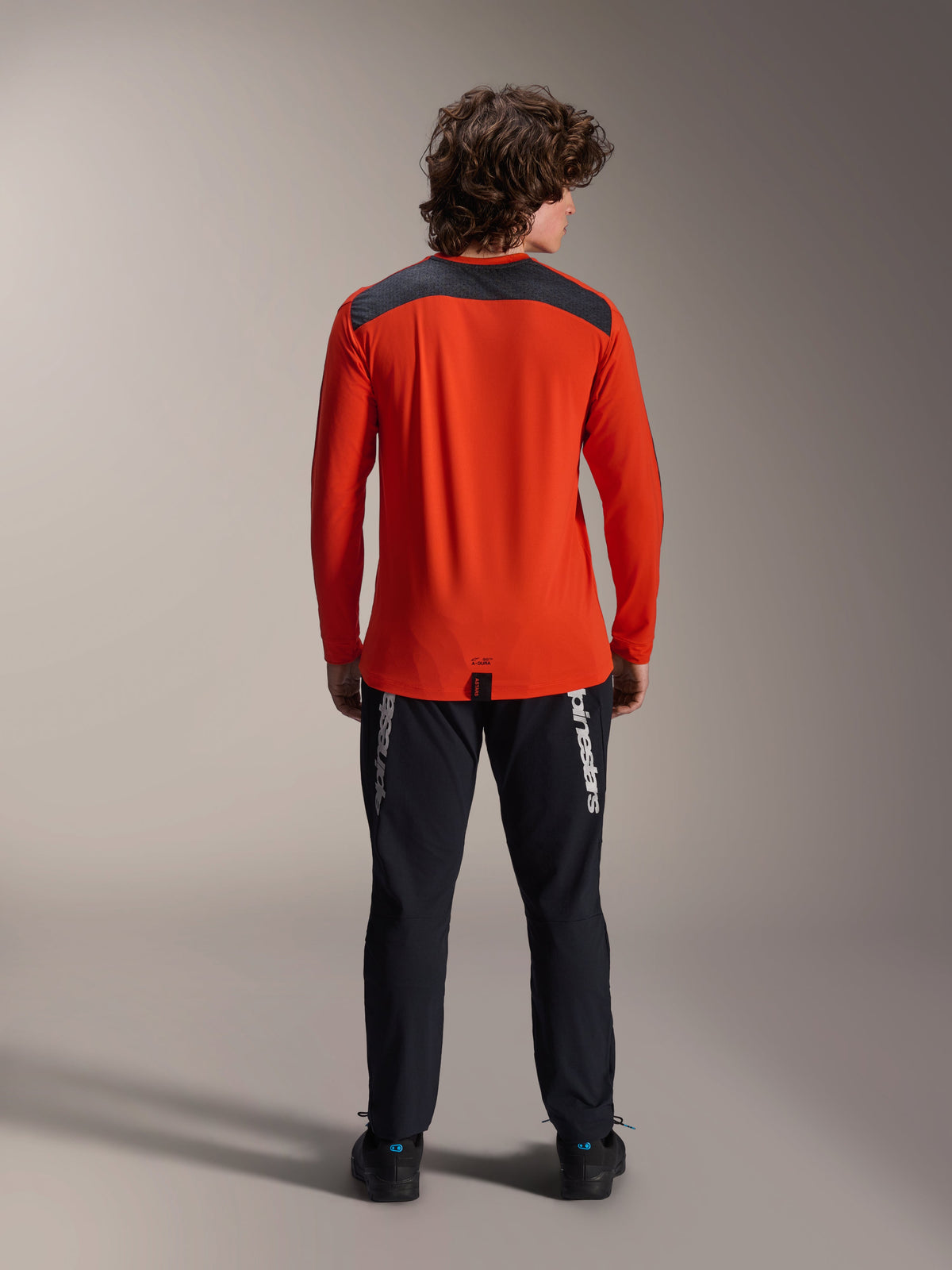 Alpinestars A-Dura Insert Astar Long Sleeve Jersey in Spicy Orange, back view showing charcoal gray shoulder panel, worn with black Alpinestars pants and black shoes, professional studio shot for MTB performance gear