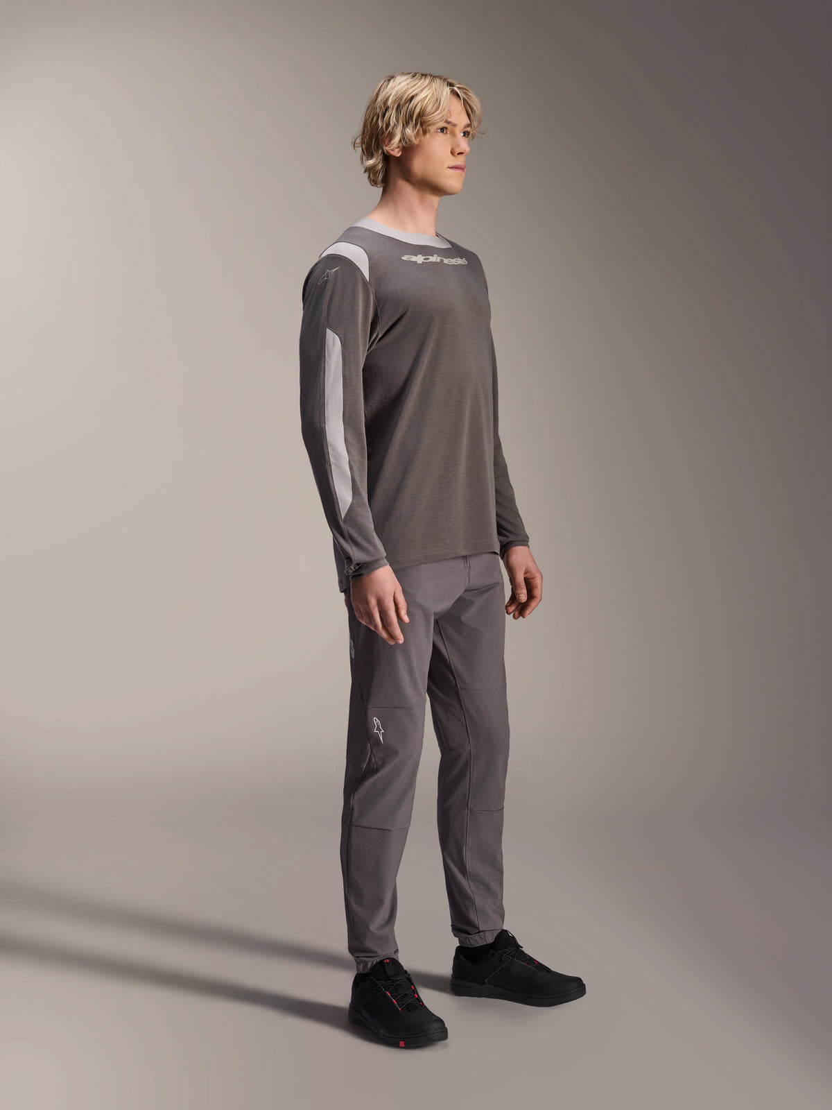 Male model wearing Alpinestars A-Dura Dri Wool Long Sleeve Jersey in Dark Gray, paired with matching gray pants and black shoes, featuring light gray mesh panels on the shoulders and sleeves, technical MTB gear for off-road cycling
