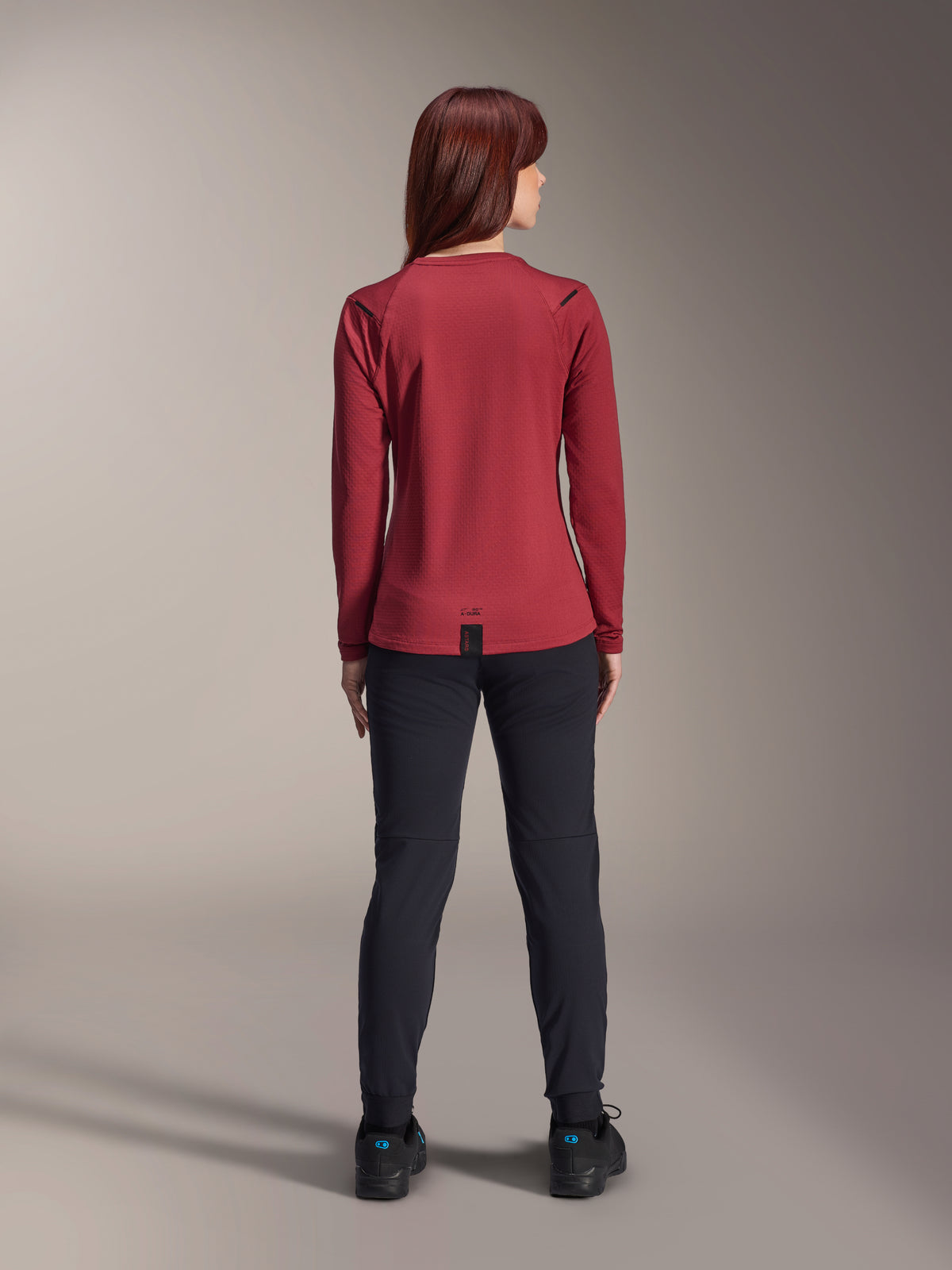 Woman wearing Alpinestars Stella A-Dura Thermal Jersey, Burgundy, rear view, long-sleeve insulated cycling jersey with an extended drop tail, paired with black pants and black riding shoes, outdoor mountain bike apparel.