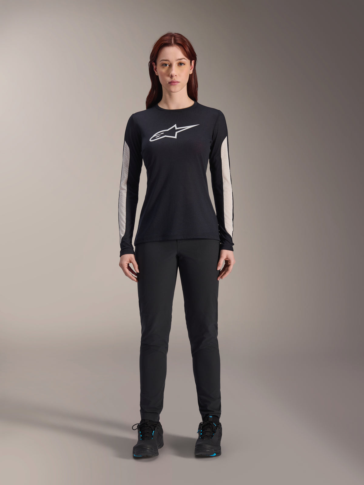 Woman wearing Alpinestars Stella A-Dura Dri Astar Long Sleeve Jersey, Black with white chest logo and light-colored mesh sleeve panels, paired with black pants and black shoes, technical mountain bike apparel