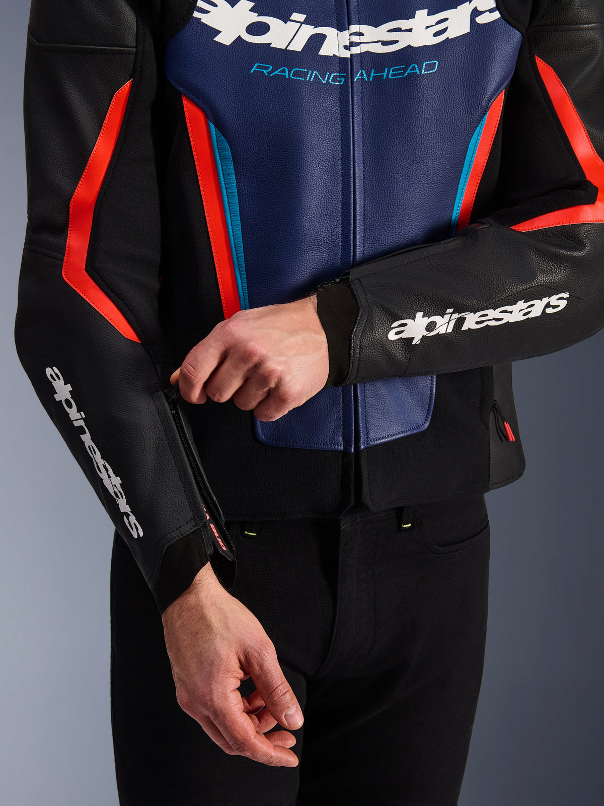 Alpinestars GP Force V2 Leather Jacket in Black, Blue, and Red Fluo, close-up view of a person adjusting the sleeve cuff zipper, featuring white Alpinestars logos on the forearm and chest, textured bovine leather construction for sport motorcycle riding