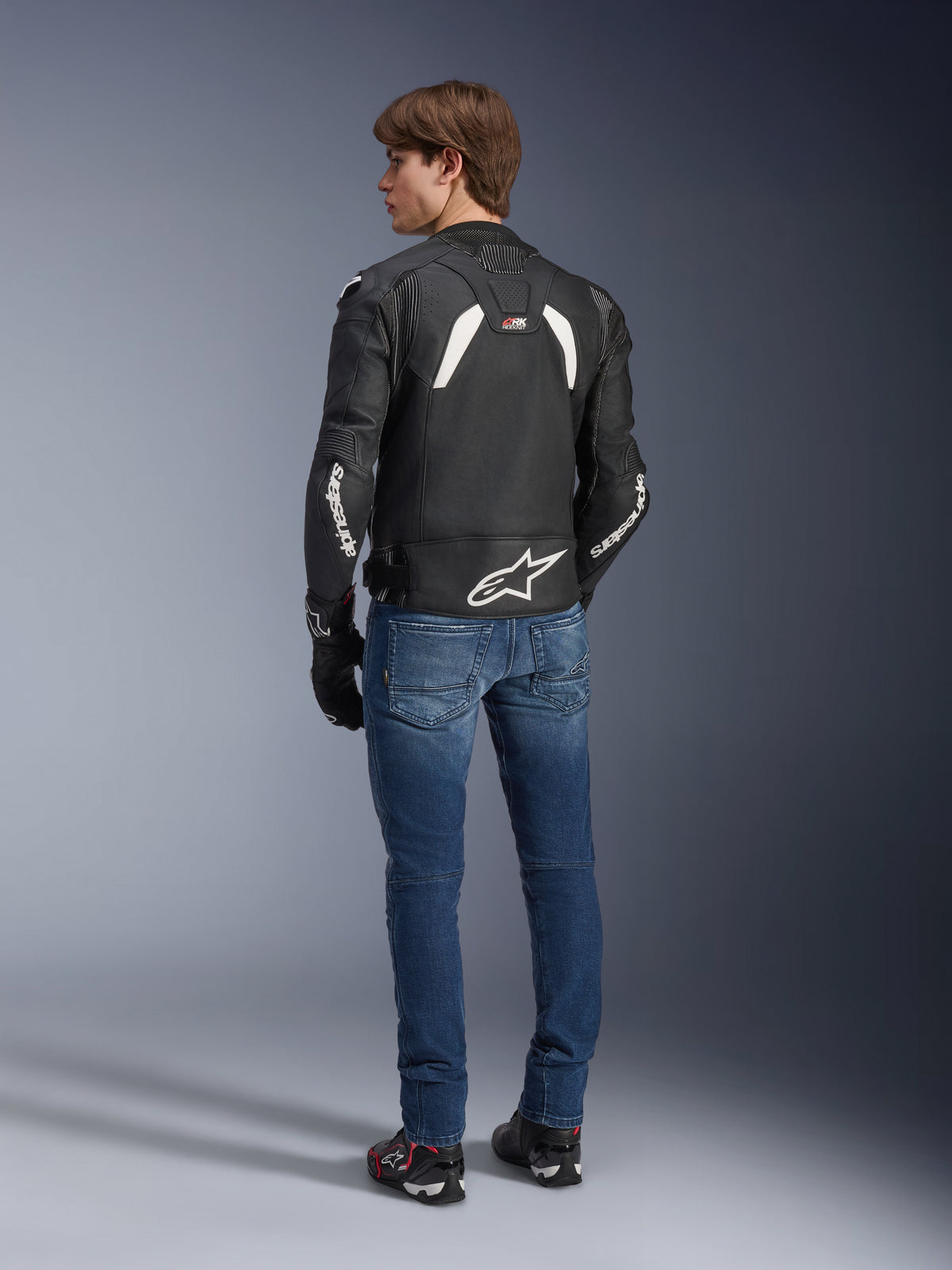Alpinestars GP Plus R V4 Rideknit Leather Jacket in Black and White, rear view on a model, featuring white Alpinestars branding on sleeves and a large star logo on the lower back, worn with blue jeans and black riding shoes, technical motorcycle gear.