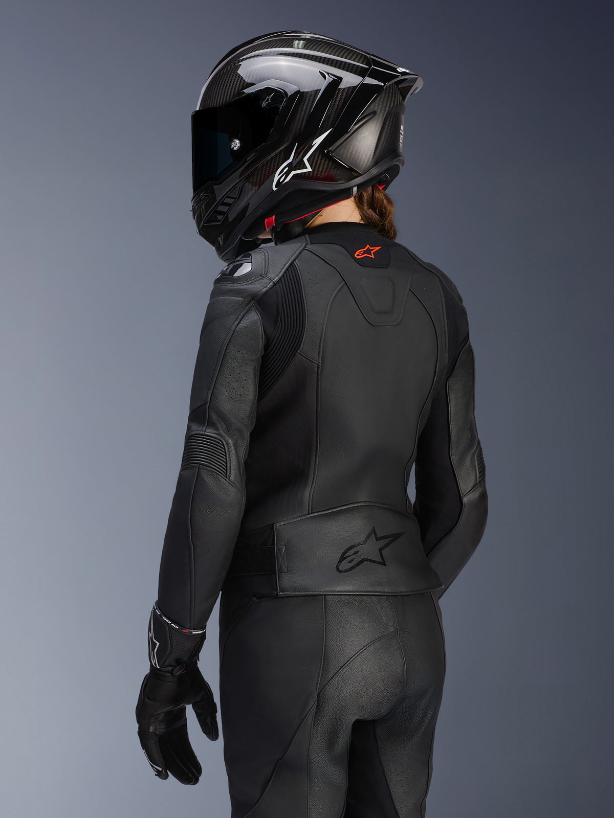 Female rider from the back wearing the Alpinestars Women's Stella GP Plus V4 Leather Jacket in Black, matching leather pants and gloves, and a black carbon-style full-face helmet, highlighting the jacket's aerodynamic fit and embossed logo on the lower back.