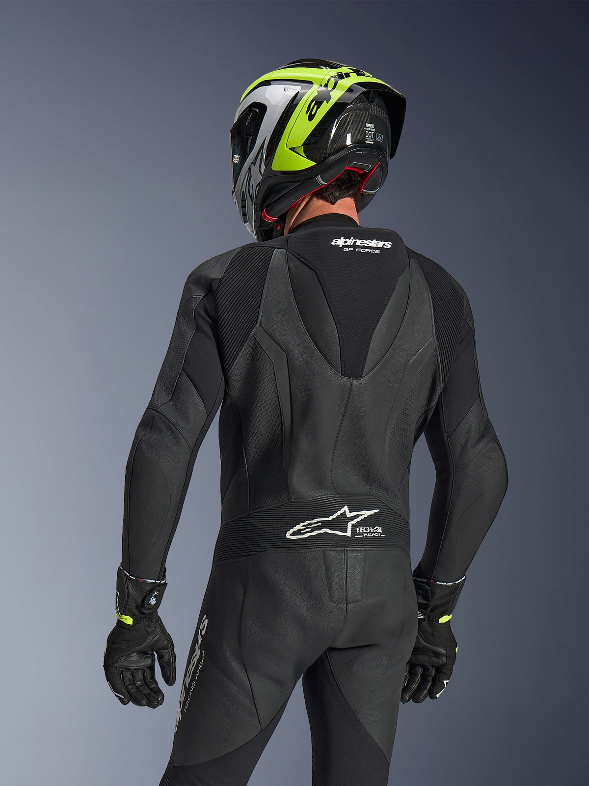 Alpinestars GP Force V2 Leather Suit 1-Piece, Black, rear view showing aerodynamic speed hump and perforated leather construction, worn with a black and neon yellow racing helmet and black gloves, featuring accordion stretch panels and white Tech-Air Ready branding.