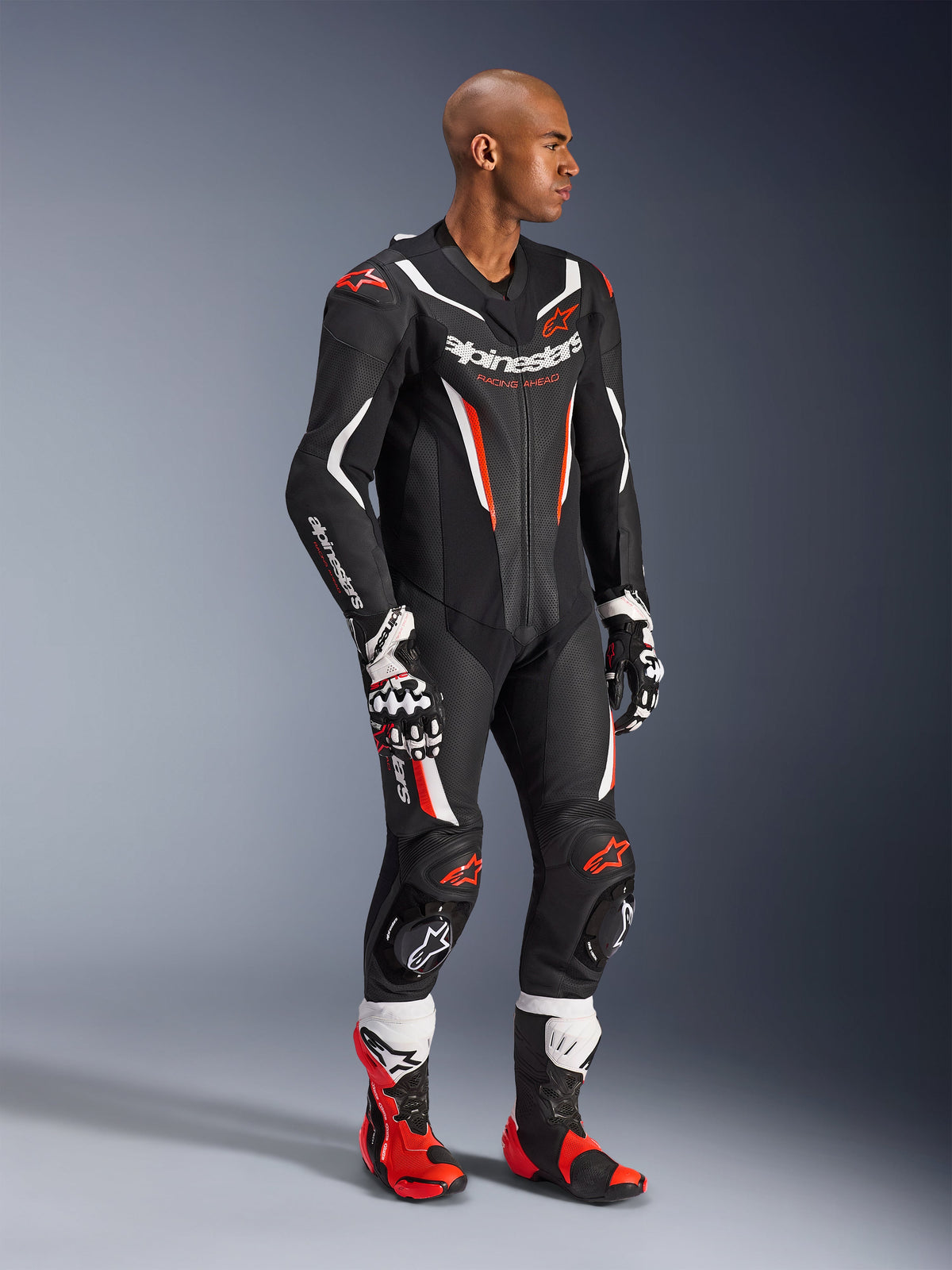 Model wearing the Alpinestars GP Force V2 Leather Suit 1-Piece in Black White Red Fluo, high-performance racing gear with perforated leather panels and DFS Lite shields, styled with matching red and white racing boots and gloves, professional motorsport aesthetic.