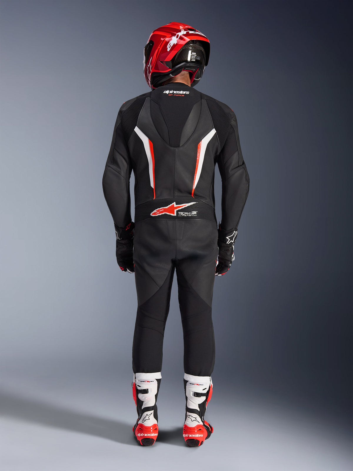 Rider viewed from behind wearing the Alpinestars GP Force V2 Leather Suit 1-Piece in Black White Red Fluo, featuring an aerodynamic speed hump and Tech-Air Ready branding, coordinated with a red racing helmet and white and red boots, professional track gear design.