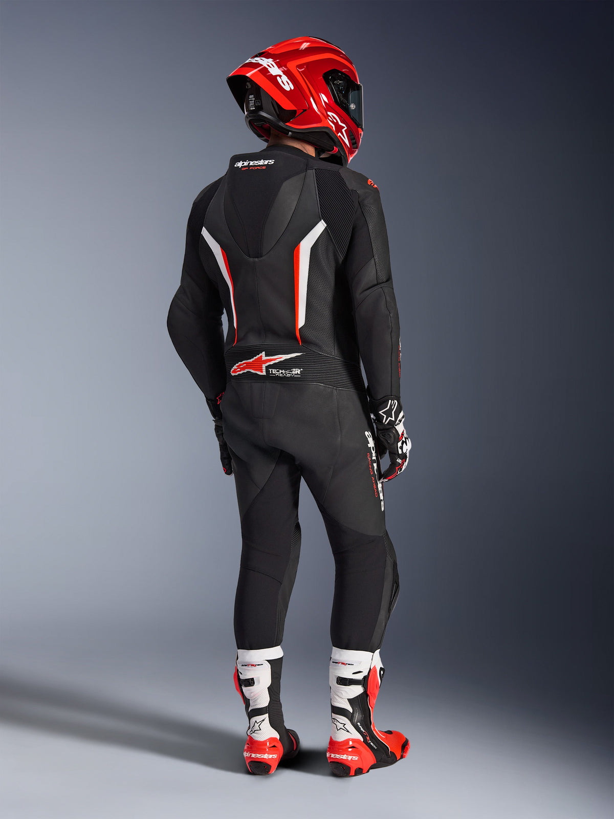 Rider seen from behind wearing the Alpinestars GP Force V2 1-Piece Leather Suit in Black, White, and Red Fluo, featuring an aerodynamic hump and perforated leather panels, paired with a red Alpinestars helmet, racing gloves, and red and white motorcycle boots, professional track racing gear.