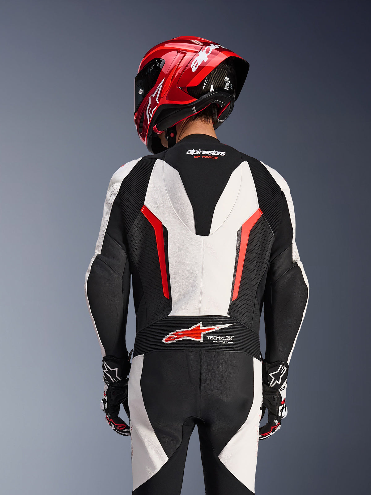 Rear view of Alpinestars GP Force V2 1-Piece Leather Suit in White Black and Red Fluo, worn with a red racing helmet, featuring Tech-Air Ready branding on the lower back, perforated leather panels, and accordion stretch zones for racing performance.