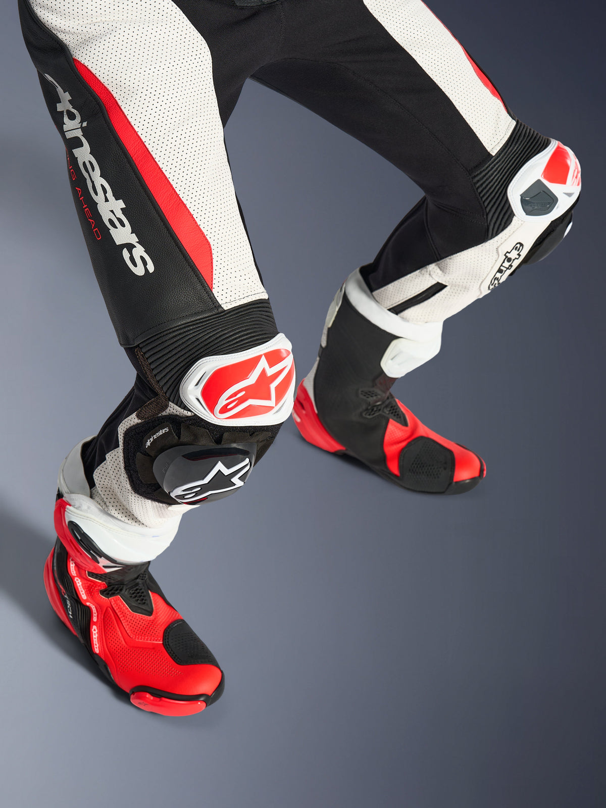 Alpinestars Woman Stella GP-R7 2-Piece Leather Suit, lower legs and racing boots, Black White and Red Fluo, featuring perforated leather, bold Alpinestars logo on the thigh, accordion stretch panels, and red-white knee sliders with matching red and black technical boots.