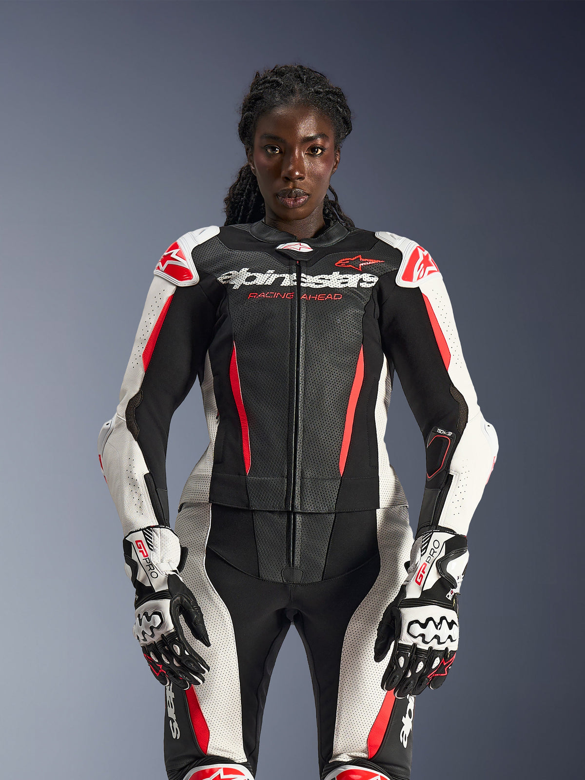 Female model wearing the Alpinestars Stella GP-R7 2-Piece Leather Suit in Black, White, and Red Fluo, featuring perforated leather panels and matching GP Pro gloves, anatomically optimized for women riders.