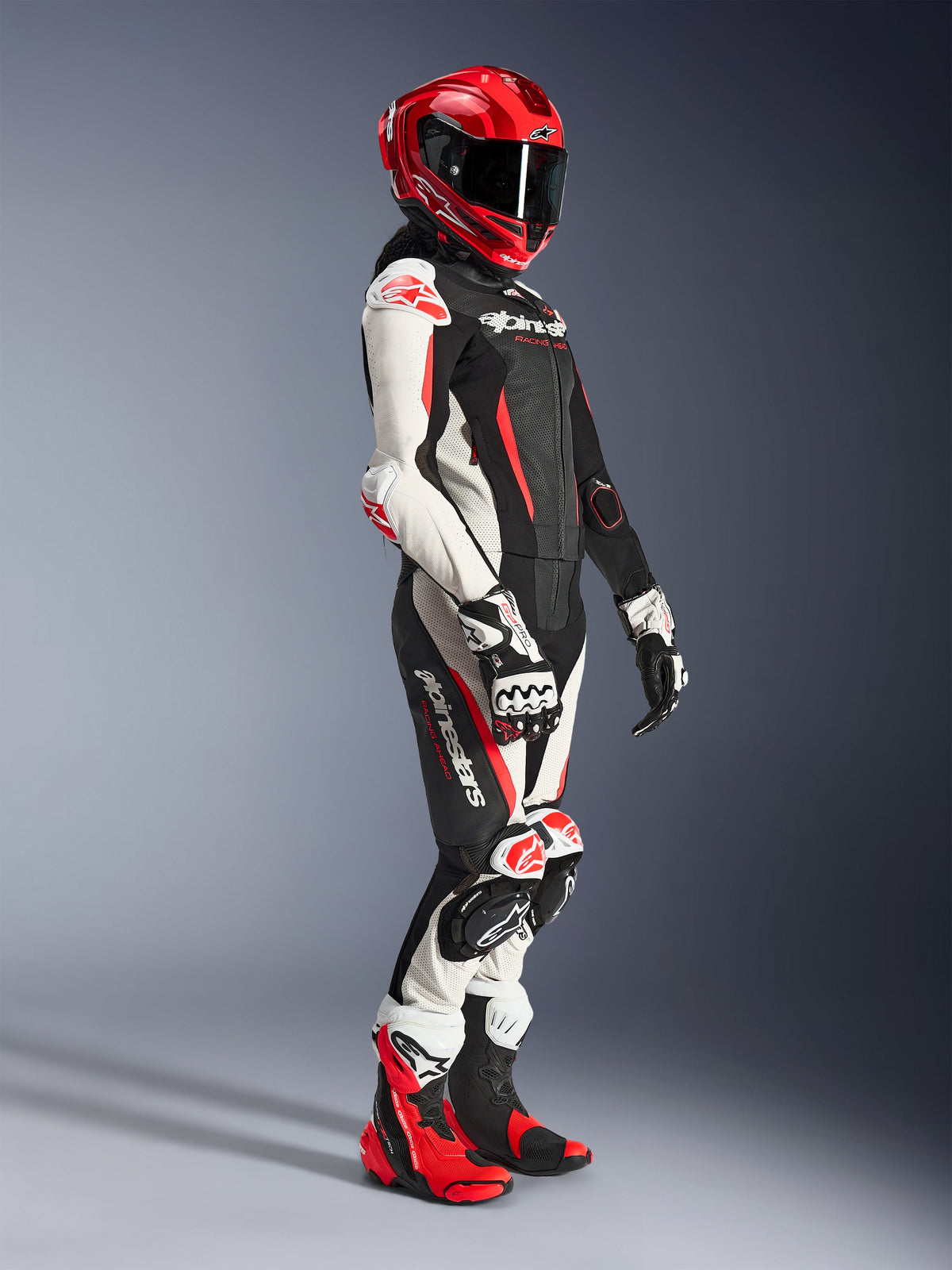 Woman rider wearing the Alpinestars Stella GP-R7 2-Piece Leather Suit in Black, White, and Red Fluo, styled with a red full-face racing helmet, matching gloves, and performance boots, professional motorcycle racing gear for women.