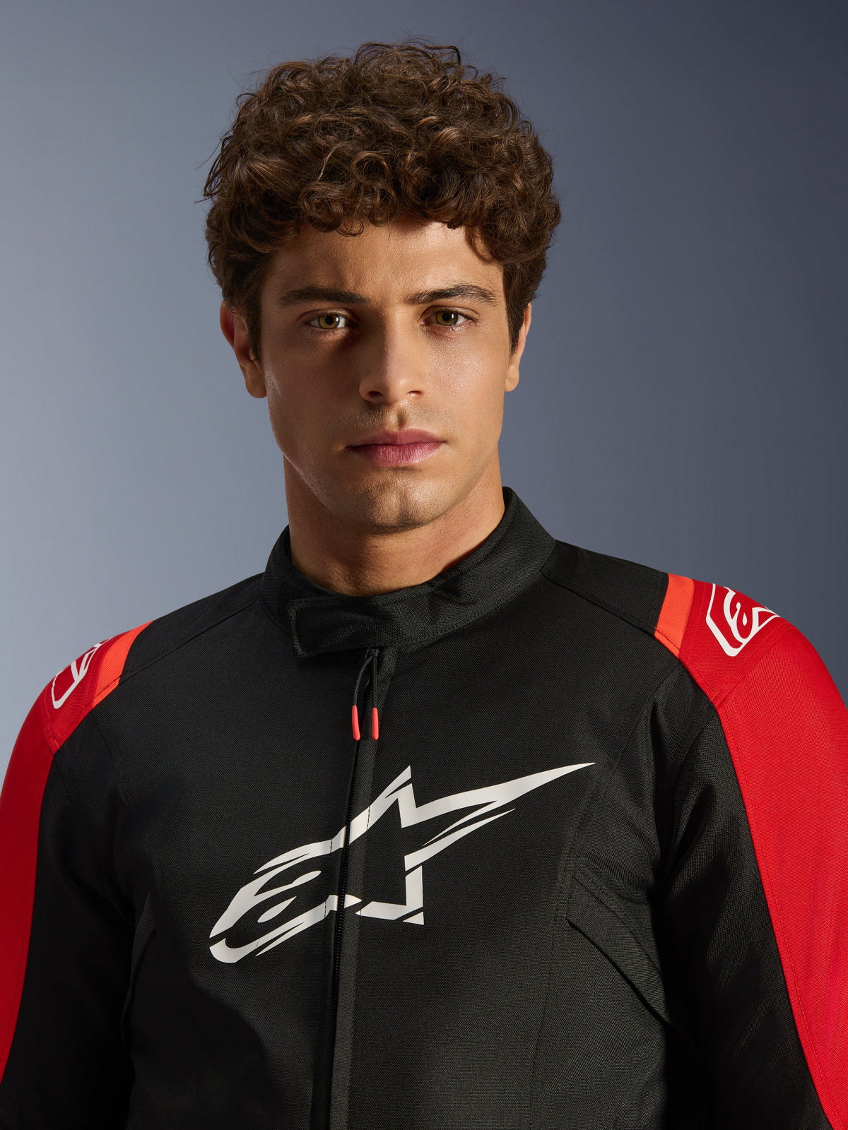 Male model wearing the Alpinestars T-SPS V2 Waterproof Jacket, Black with Red accents and white logo, close-up portrait view, textile sport motorcycle jacket