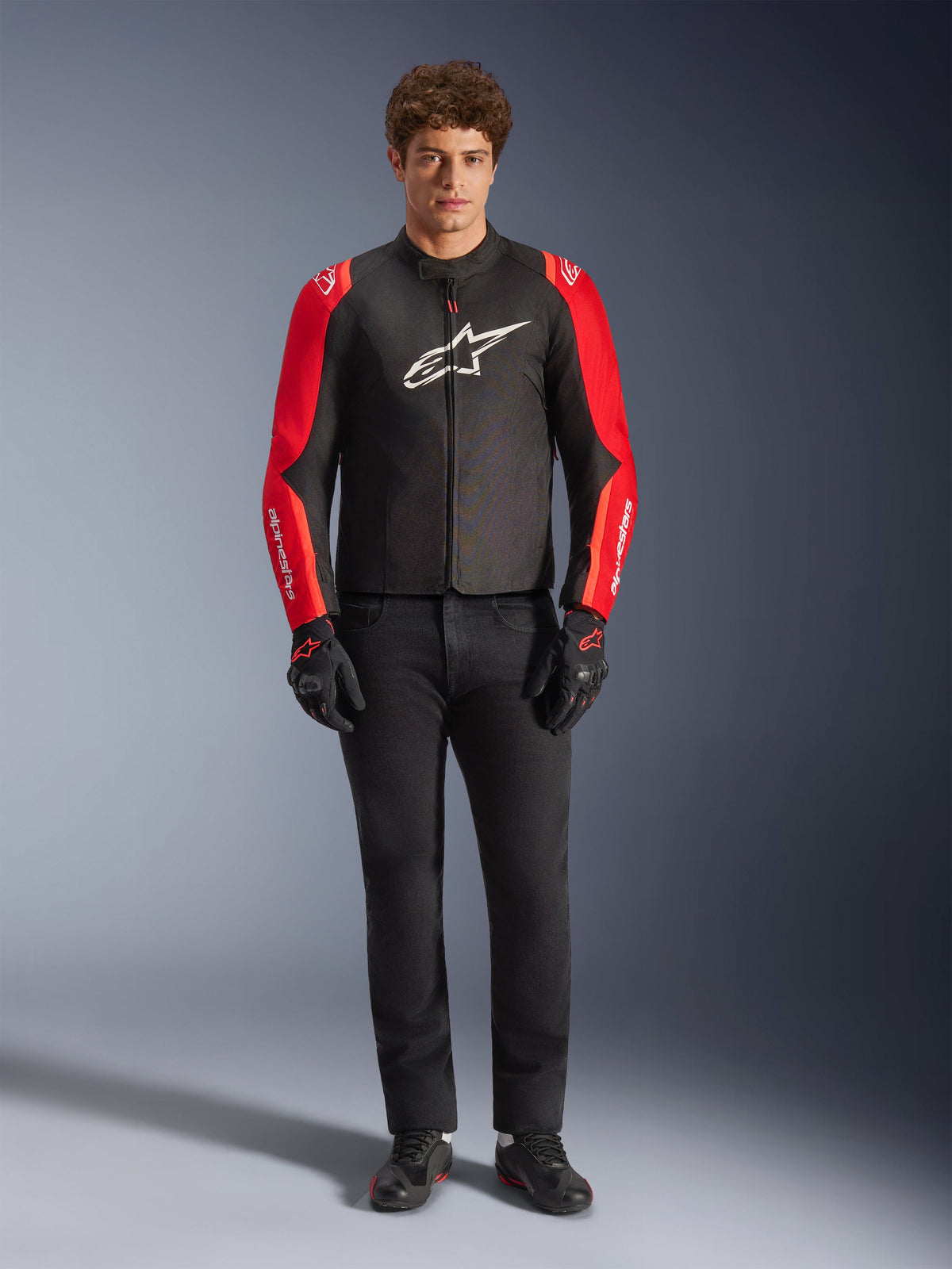 Male model wearing the Alpinestars T-SPS V2 Waterproof Jacket, Black with Red and Red Fluo sleeves, white logo on chest, paired with black pants, black riding gloves and shoes, sporty motorcycle road gear design