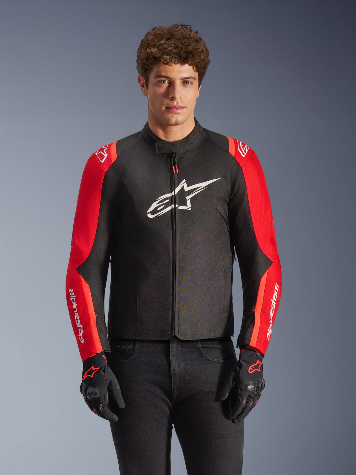Alpinestars T-SPS V2 Waterproof Jacket, textile motorcycle jacket, Black and Red, worn by a male model with matching black gloves, featuring a large white logo on the chest and white branding on red sleeves, sporty silhouette for all-weather road riding