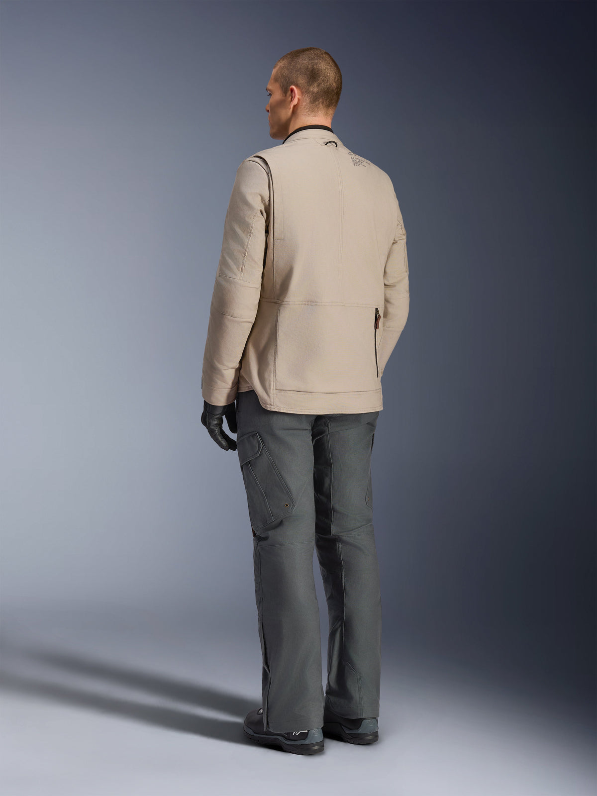 Alpinestars Borrego Drystar® Jacket in Warm Gray, rear view showing the rugged waxed-canvas texture, rear shoulder expansion panels, and large zippered back pocket, worn by a model with charcoal gray adventure pants and black gloves.