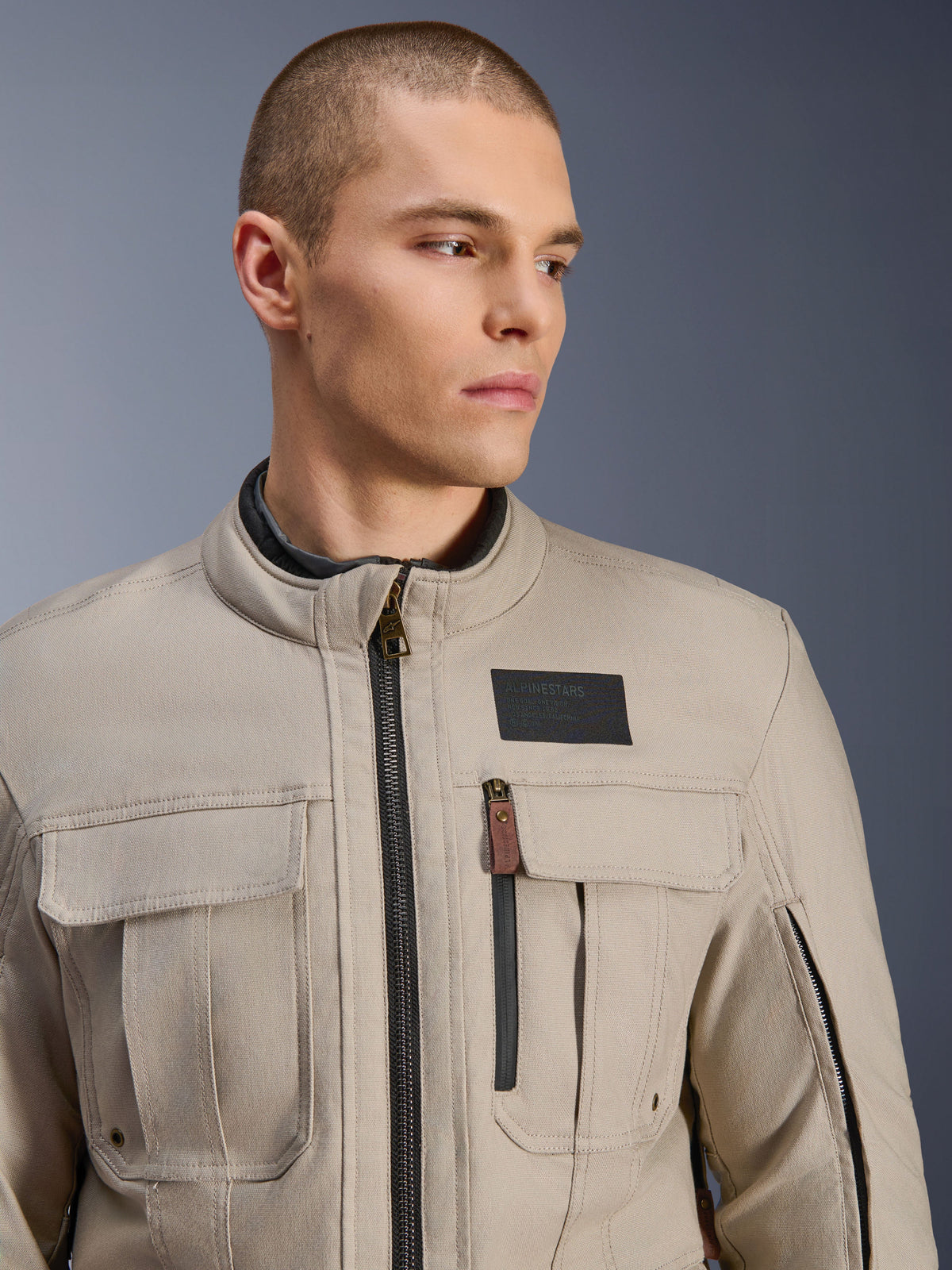 Male model wearing the Alpinestars Borrego Drystar® Jacket in Warm Gray, adventure motorcycle jacket with a classic waxed-canvas look, functional chest cargo pockets, and a black Alpinestars logo patch.