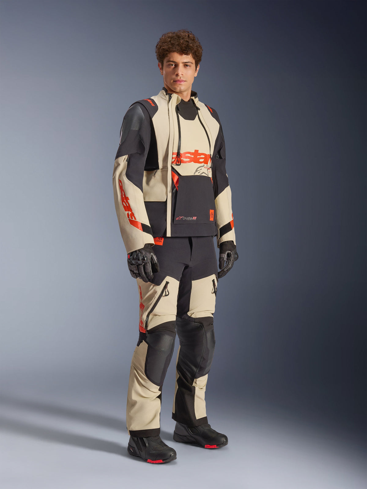 Male model wearing the Alpinestars Halo Pro Drystar® XF Laminated Jacket in Mastic Black, featuring beige and black panels with red accents, paired with matching adventure pants and black gloves, designed for all-terrain adventure motorcycle riding.