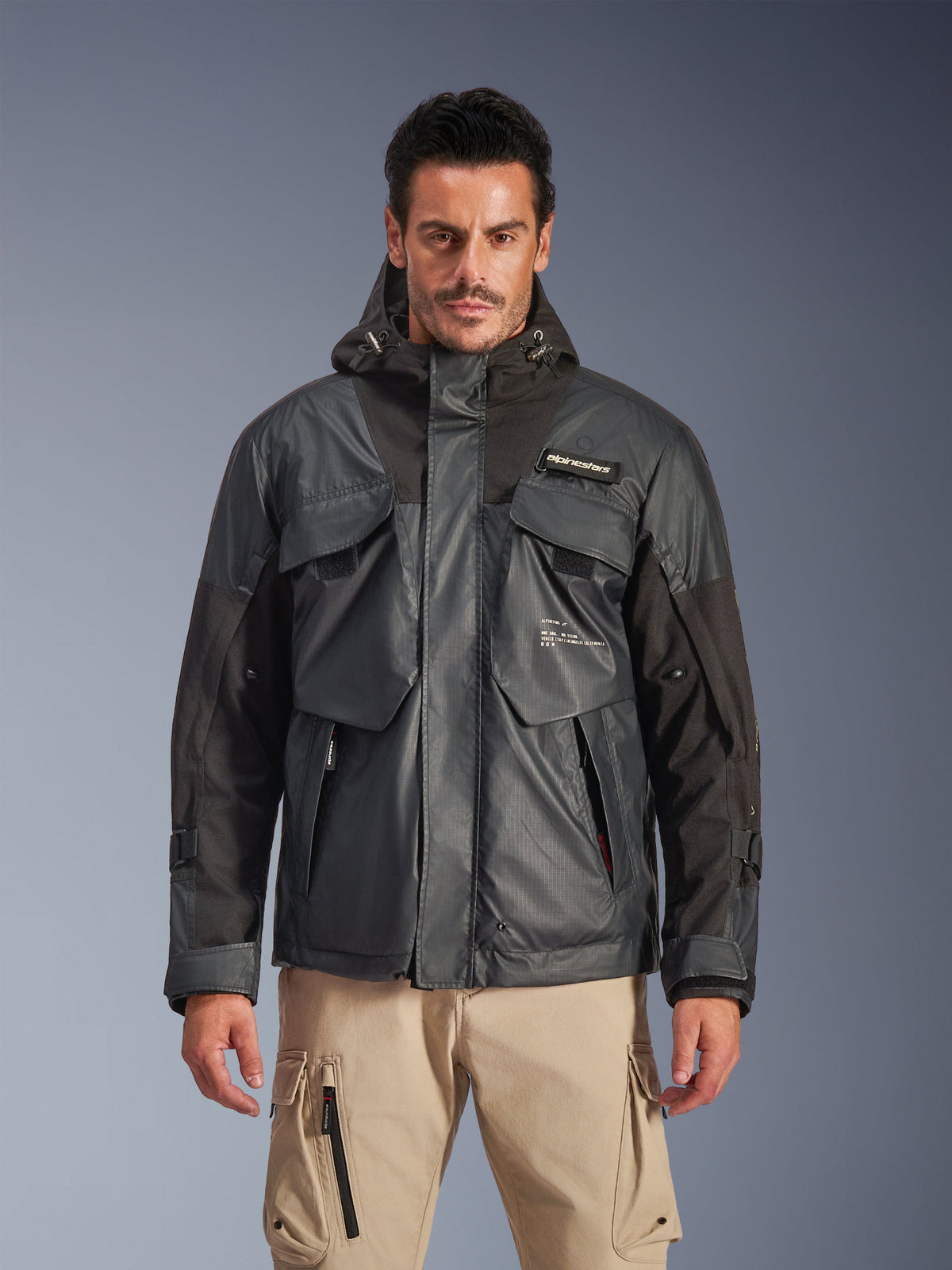 Alpinestars Mospher Waterproof Jacket, Men's Urban Motorcycle Parka, Black and Dark Gray ripstop fabric, hooded design with multiple large utility pockets, worn by a model with khaki cargo pants for a tactical look