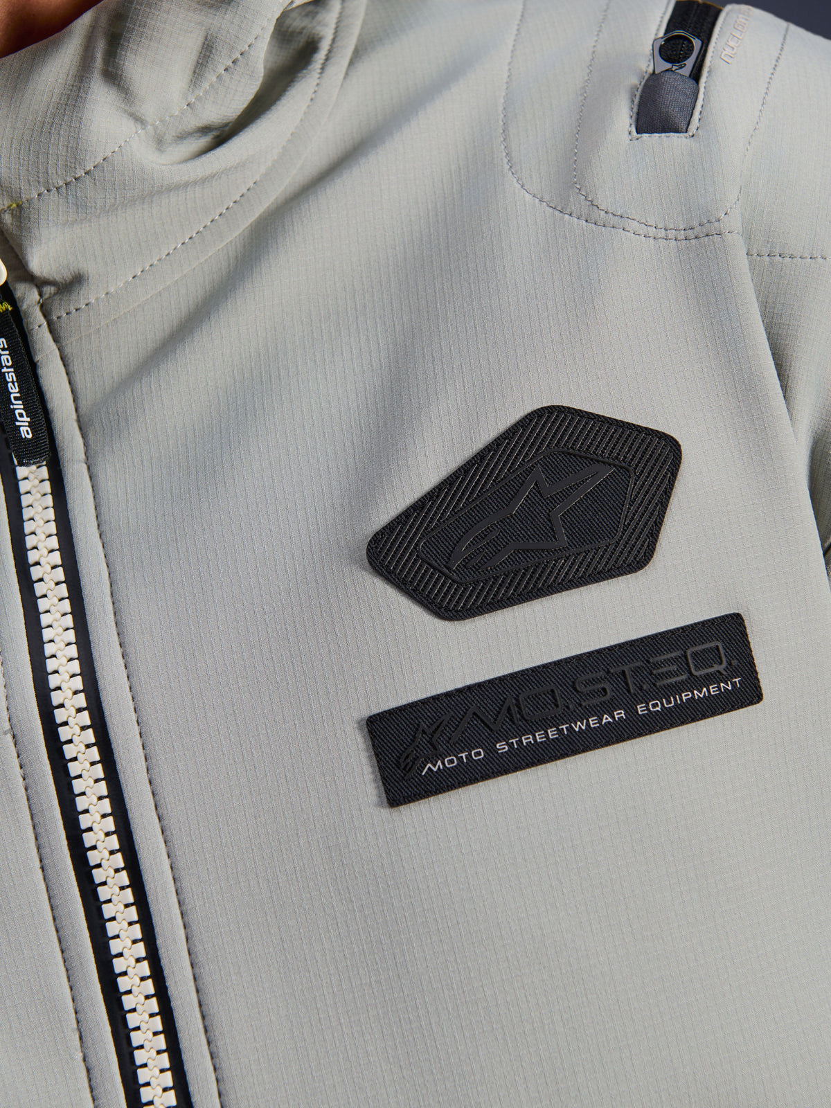 Alpinestars Moblast Waterproof Jacket, Iron Gray, close-up detail of chest patches featuring the Alpinestars logo and MO.ST.EQ. Moto Streetwear Equipment branding, technical ripstop fabric with heavy-duty front zipper and shoulder pocket detail.