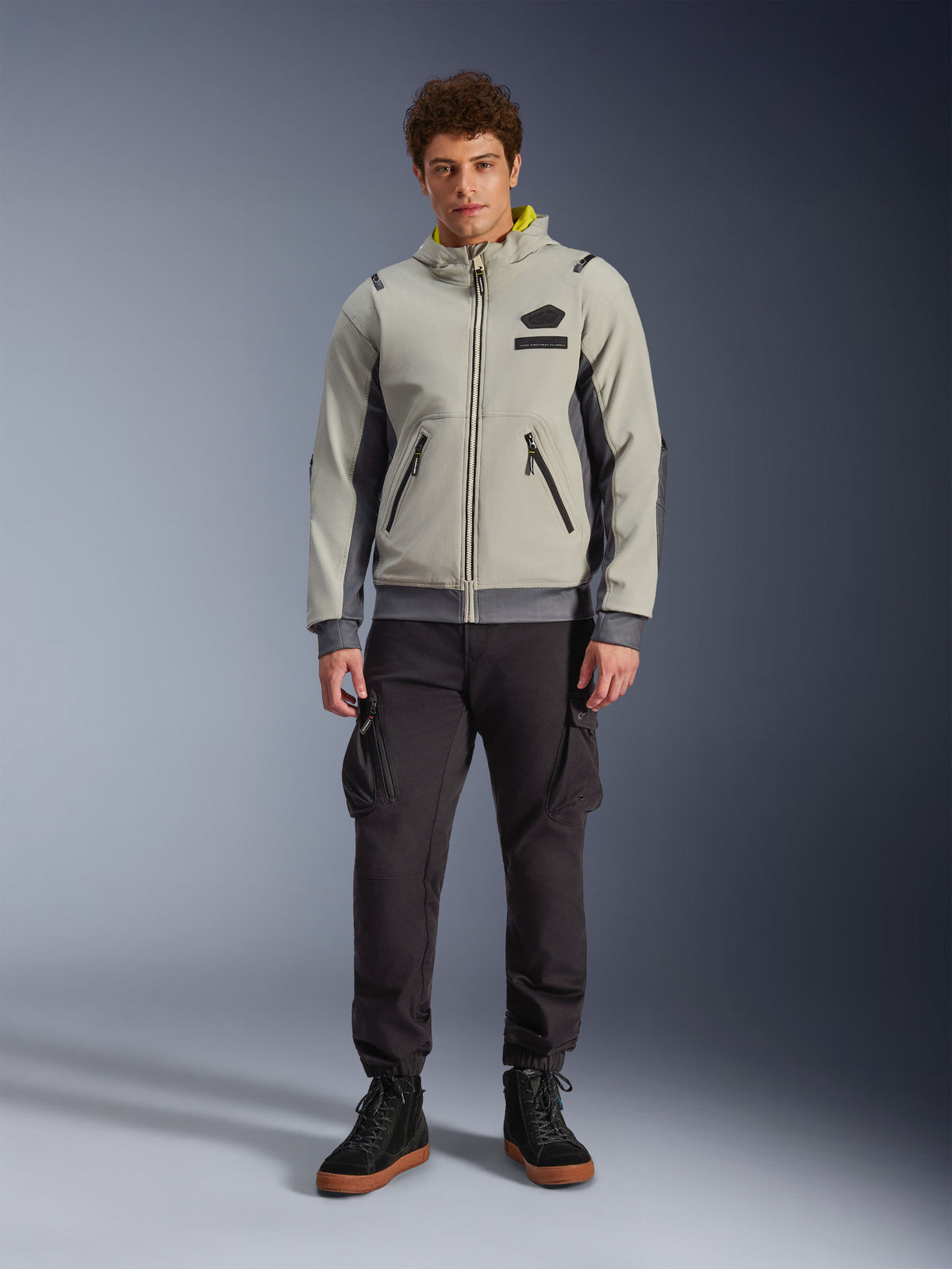 Male model wearing Alpinestars Moblast Waterproof Jacket in Iron Gray and Dark Sage, hooded motorcycle jacket with a modern technical design, paired with dark cargo riding pants and black boots, urban contemporary motorsport apparel