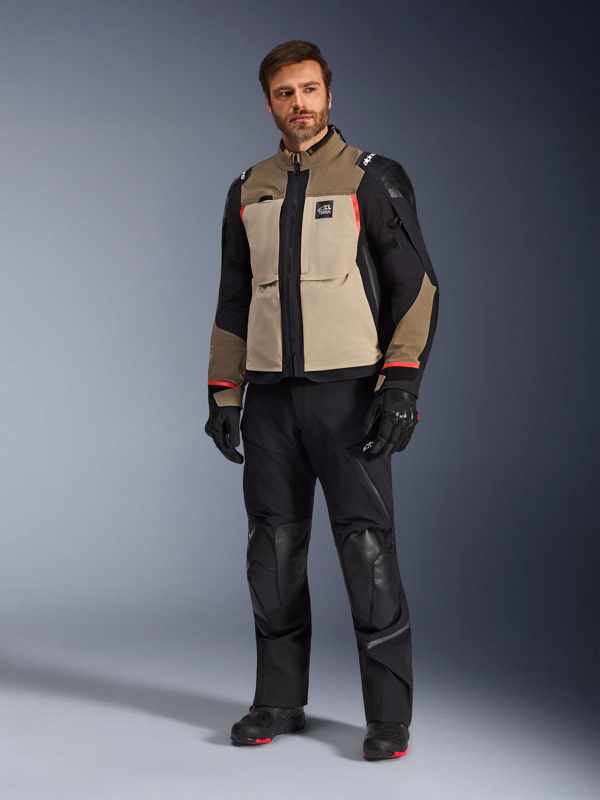 A male model wearing the Alpinestars Andes Pro Drystar® XF Jacket in Walnut Aluminum, paired with black motorcycle pants, gloves, and boots, featuring a tan and black multi-panel design with red accents for adventure touring.