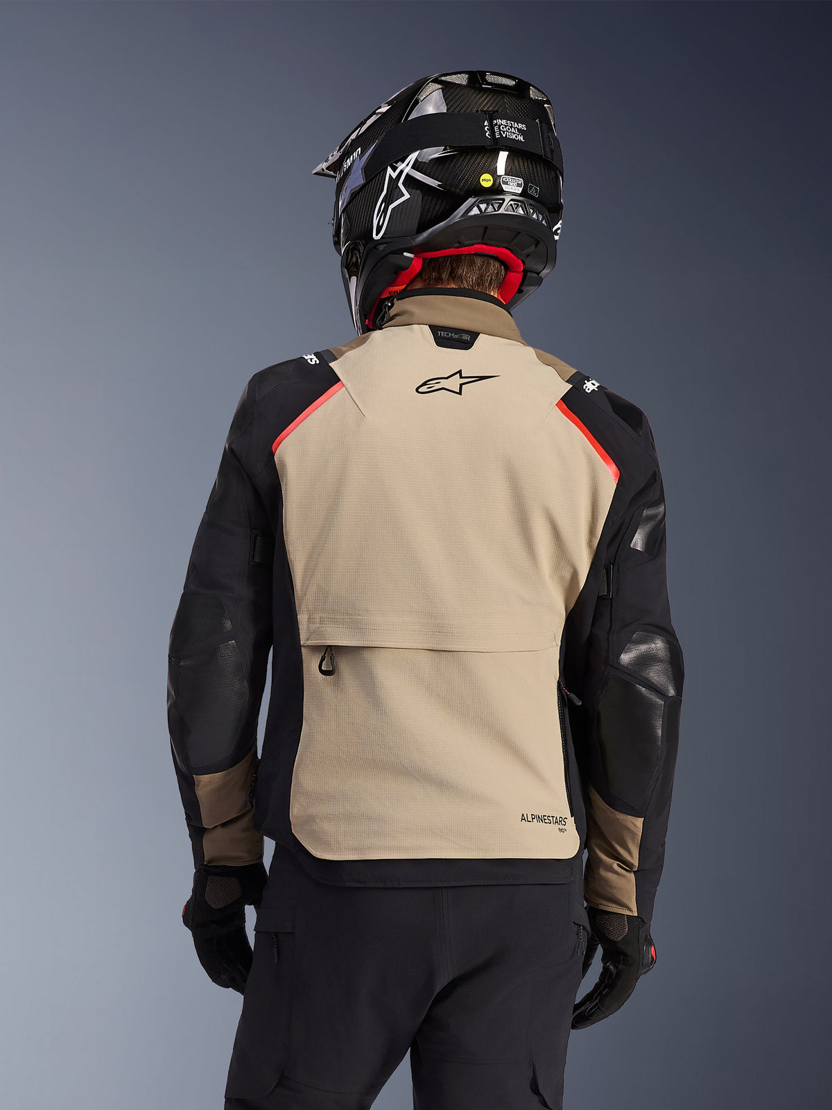 Alpinestars Andes Pro Drystar® XF Jacket in Walnut Aluminum, back view, tan back panel with large cargo pocket, black sleeves with reinforced panels, Alpinestars logo on the upper back, paired with a black helmet and gloves for adventure touring.