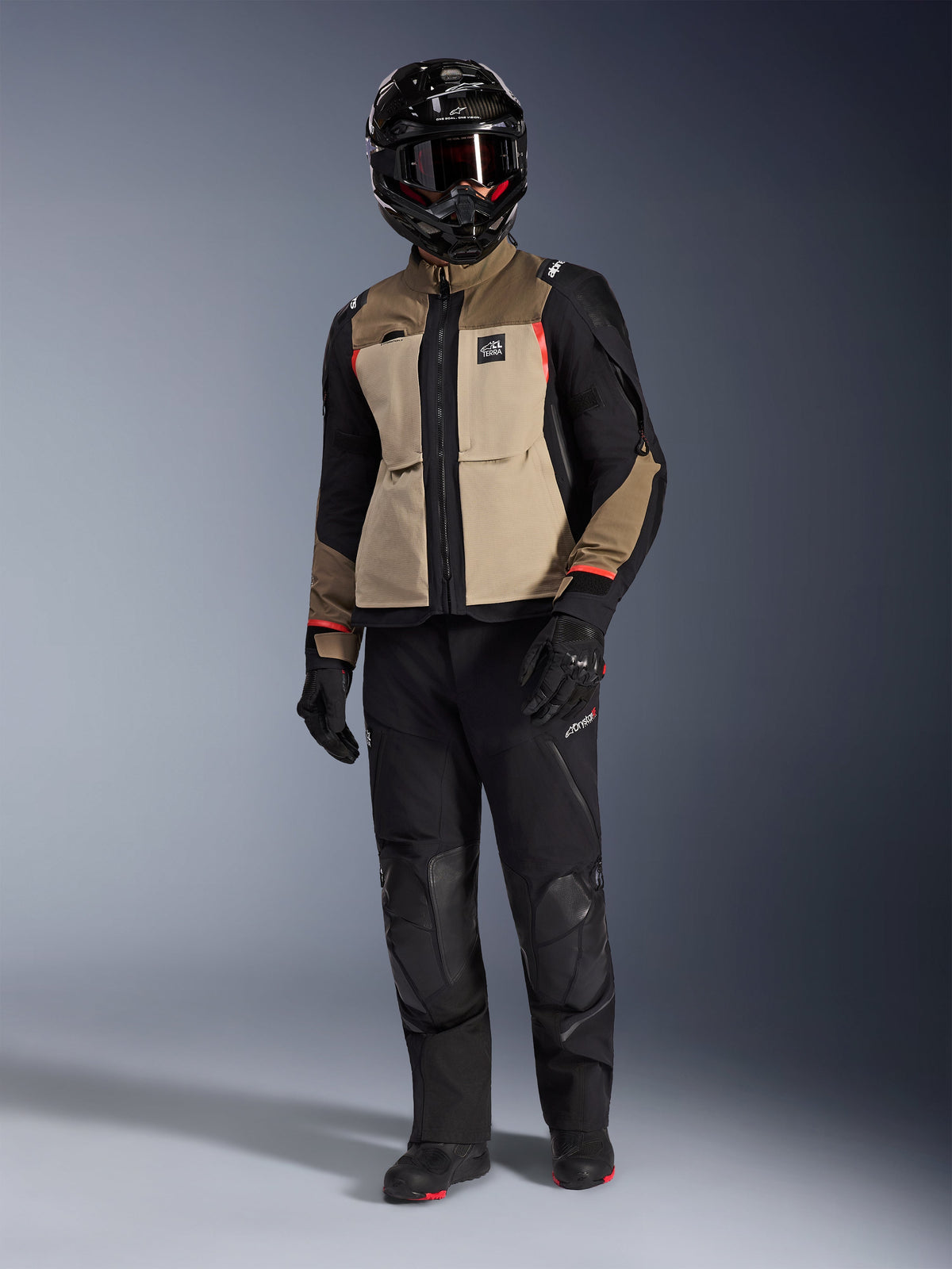 Alpinestars Andes Pro Drystar® XF Jacket in Walnut Aluminum, adventure motorcycle jacket worn with matching black pants, a black adventure helmet with goggles, and black boots, featuring tan and black panels with red accents.