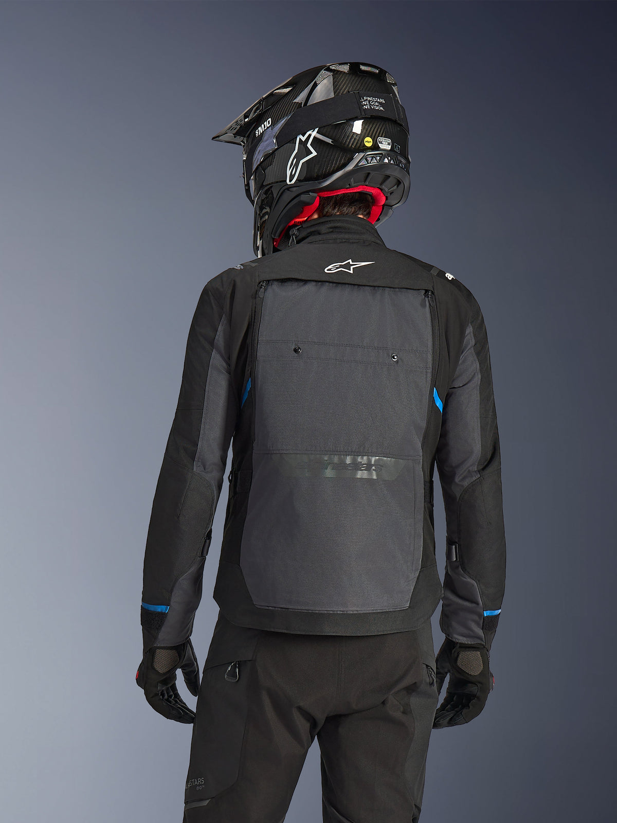Rider from behind wearing the Alpinestars Cusco Drystar® Jacket in Black and Deep Gray, featuring a large gray back ventilation panel and white logo, paired with a black off-road helmet and black riding gear for adventure touring.