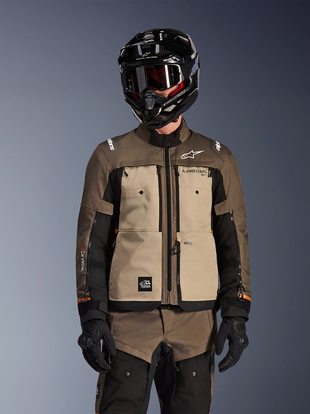 Rider wearing the Alpinestars Cusco Drystar Jacket in Walnut Aluminum Black, adventure touring motorcycle outfit featuring a black helmet with goggles and black gloves, front view showing the technical textile construction, pockets, and branding.