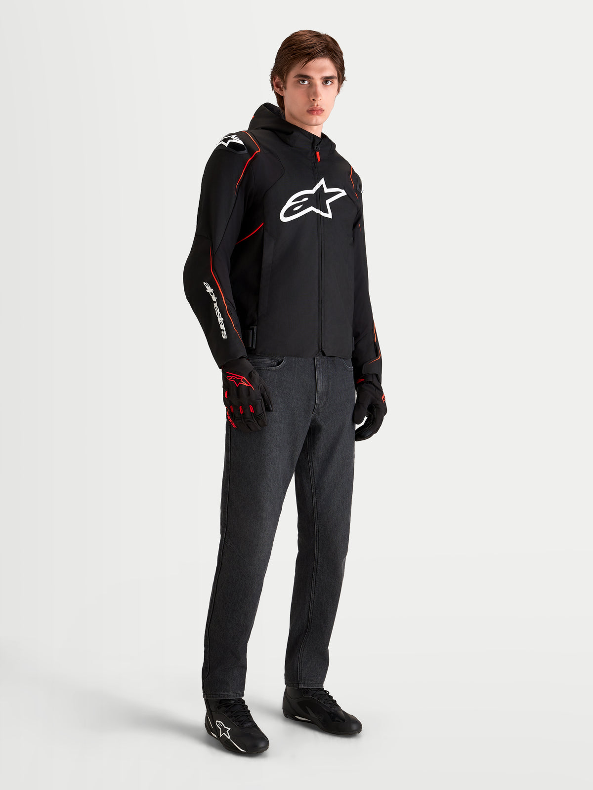 Alpinestars Zaca V2 Waterproof Jacket, men's black and red motorcycle sport jacket with hood and white chest logo, worn by a male model with matching riding gloves and dark jeans