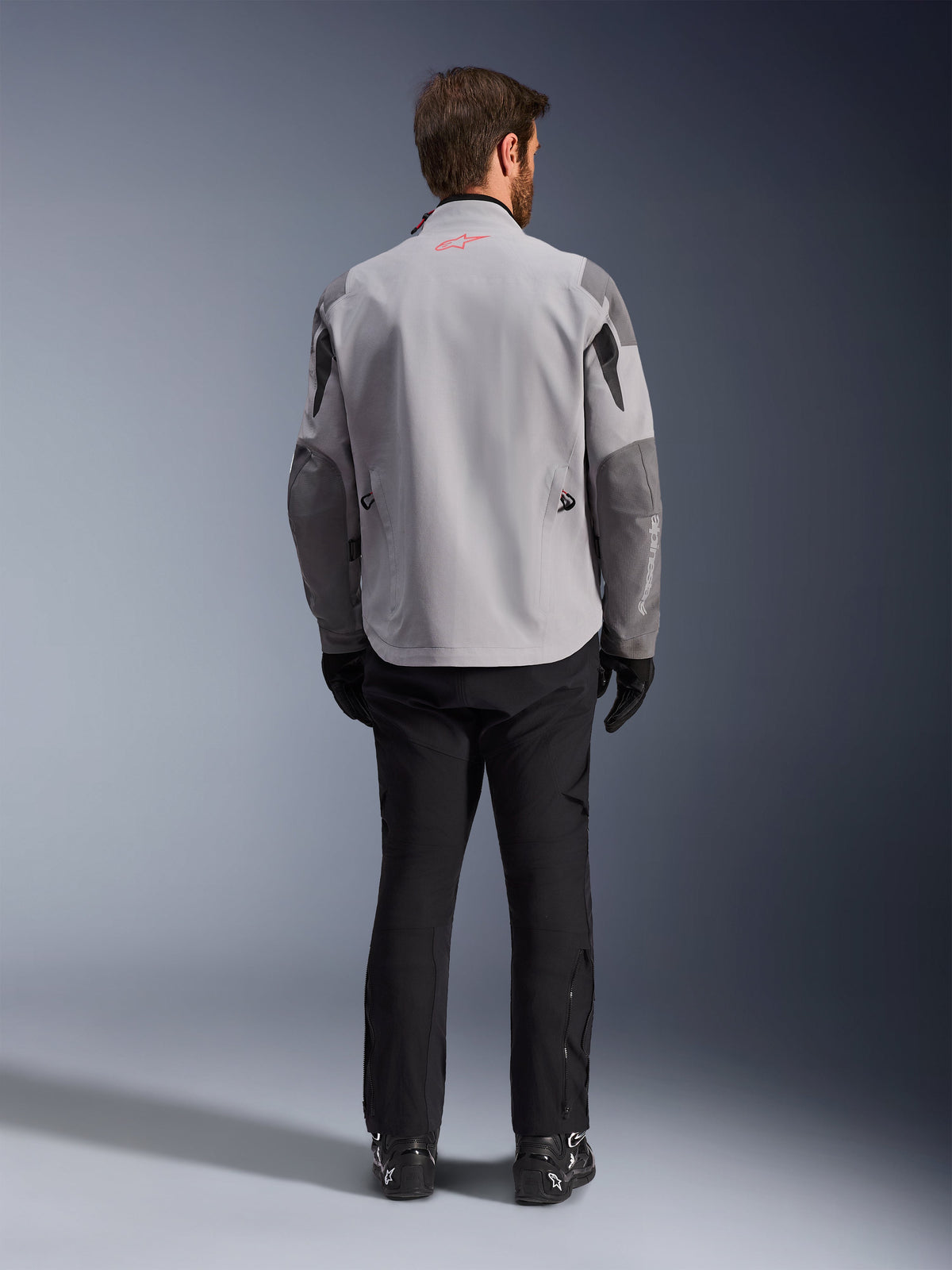 Alpinestars Maxdura Drystar®XF Laminated Armored Shell, Mid Gray and Dark Gray Motorcycle Jacket, back view, featuring a light gray body with dark gray panels on the shoulders and sleeves, small red Alpinestars star logo on the back collar, and white branding on the lower left sleeve, paired with black riding pants and boots.